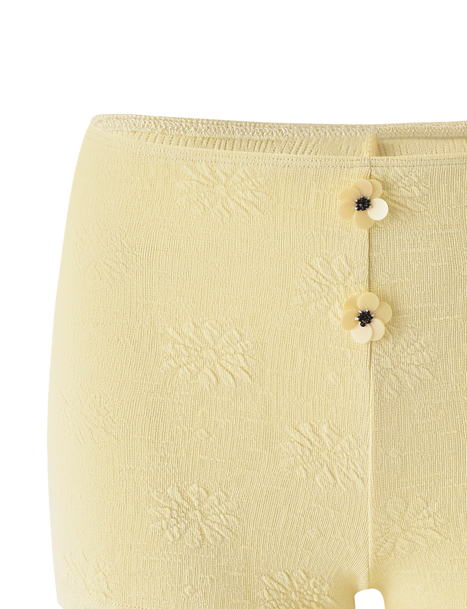 Close-up of TIGERMIST's QUINLAN SHORT - LEMON: light yellow mini shorts with a subtle floral pattern, delicate floral trim, and two small flower-shaped buttons with black centers on the front waistband.