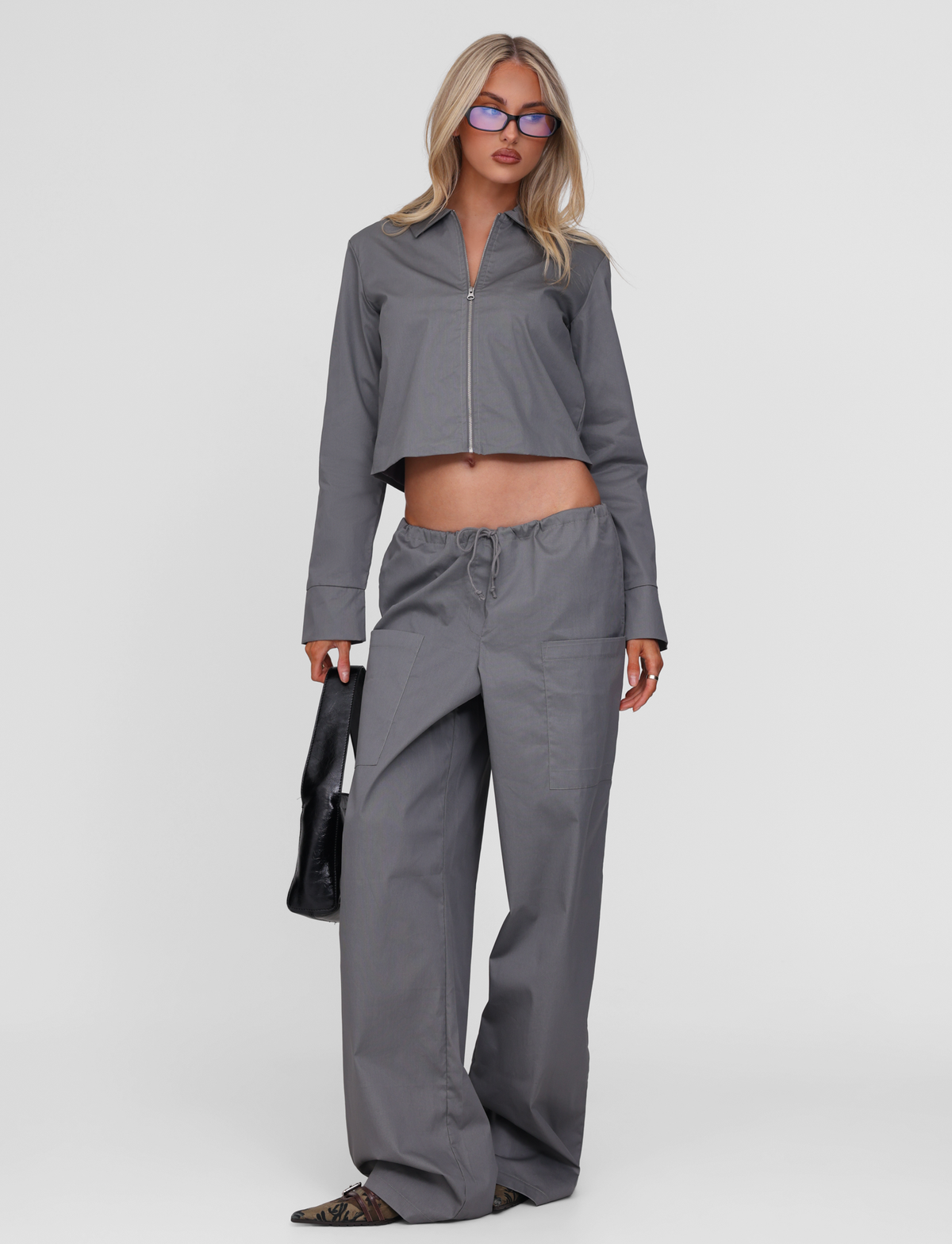 A woman with long blond hair, glasses, and pointed-toe shoes stands against a white background wearing the TIGERMIST TYSHA JACKET in charcoal with matching wide-leg pants. The boxy fit and lightly ribbed texture add subtle detail. She holds a black handbag.