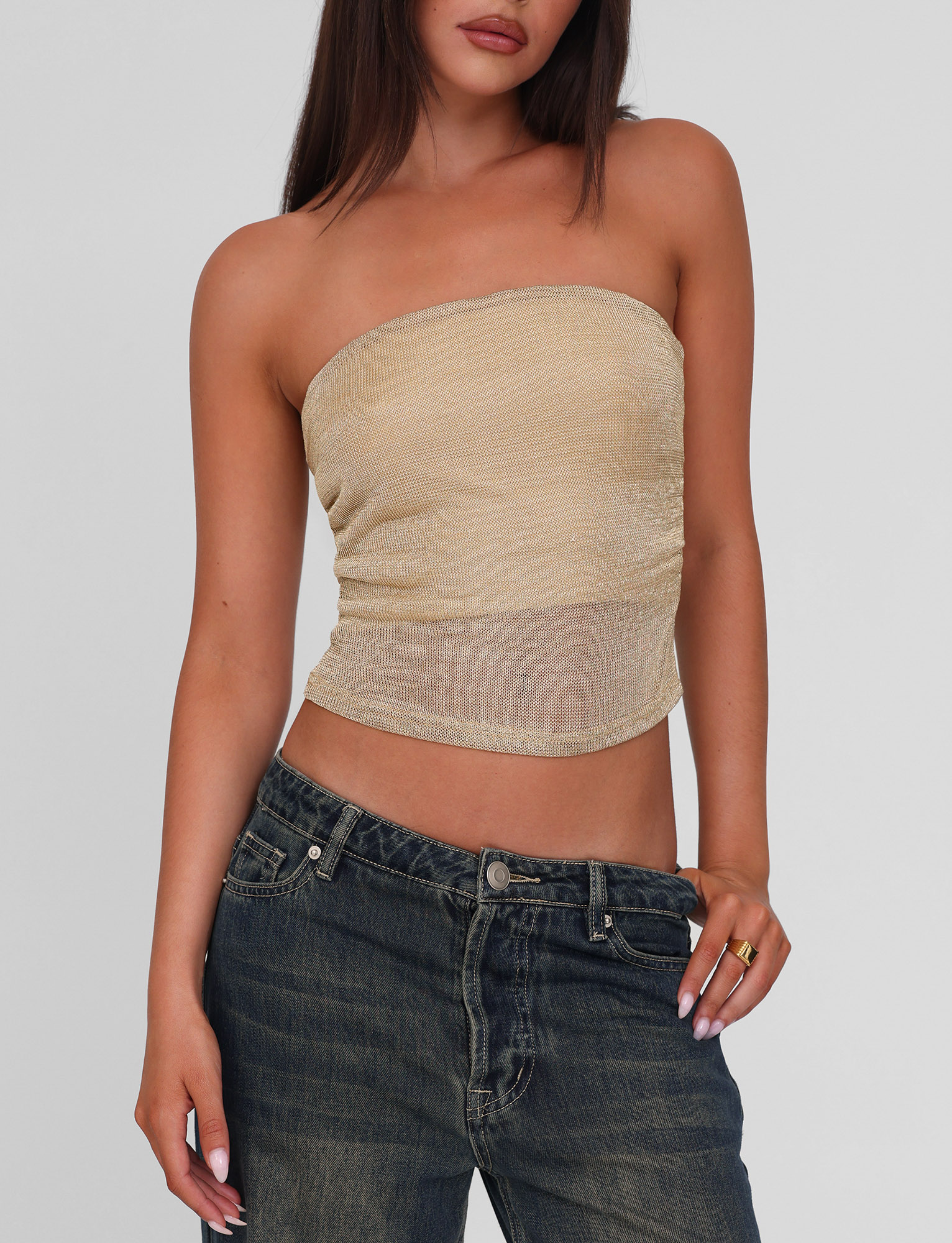 A woman models the TIGERMIST HARRIETT TUBE TOP in gold with low-rise blue jeans, posing with one hand in her pocket and the other by her side against a plain background.