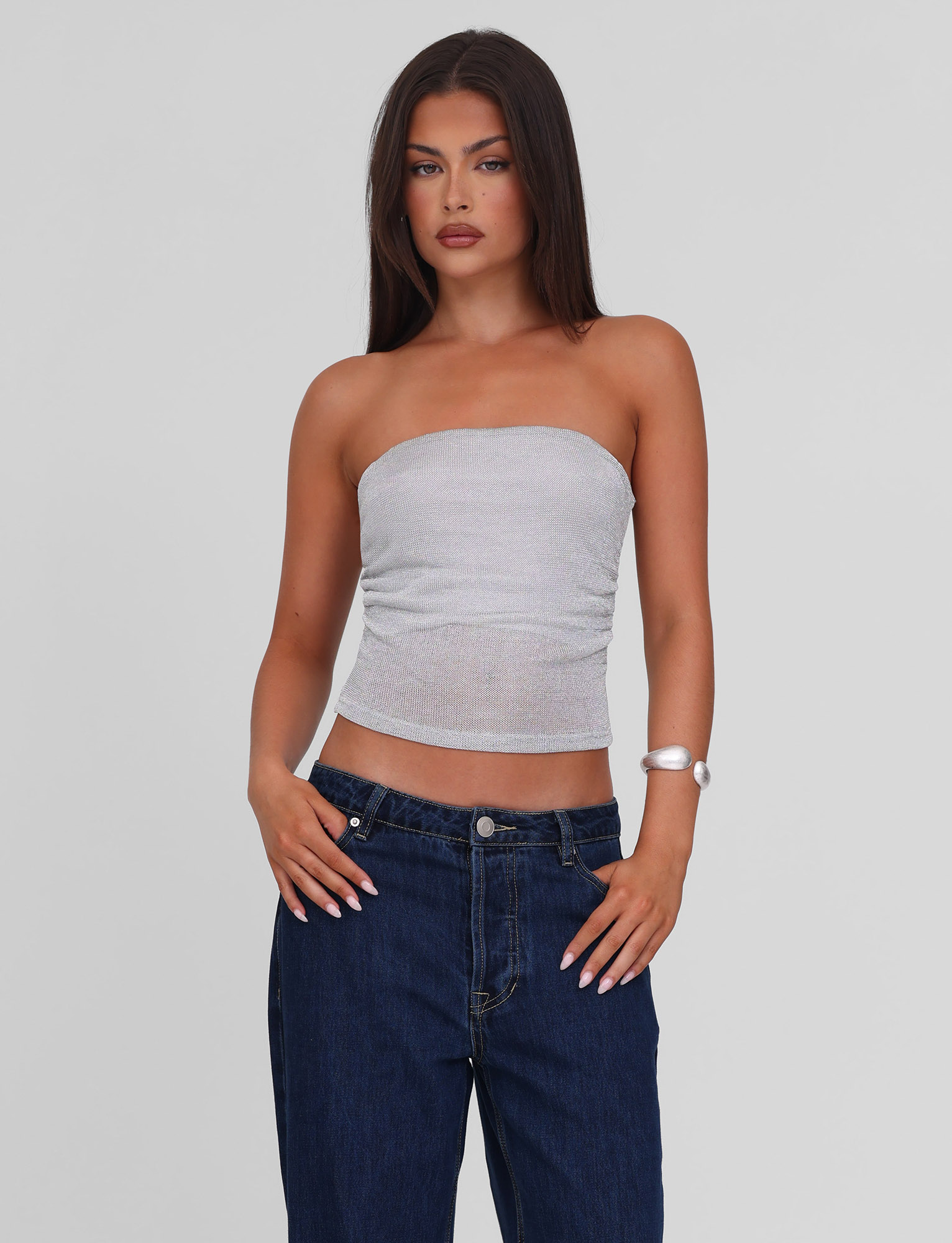 A woman with long brown hair wears the TIGERMIST HARRIETT TUBE TOP in silver sparkle jersey and dark blue jeans, standing against a plain light gray background with one hand on her hip.
