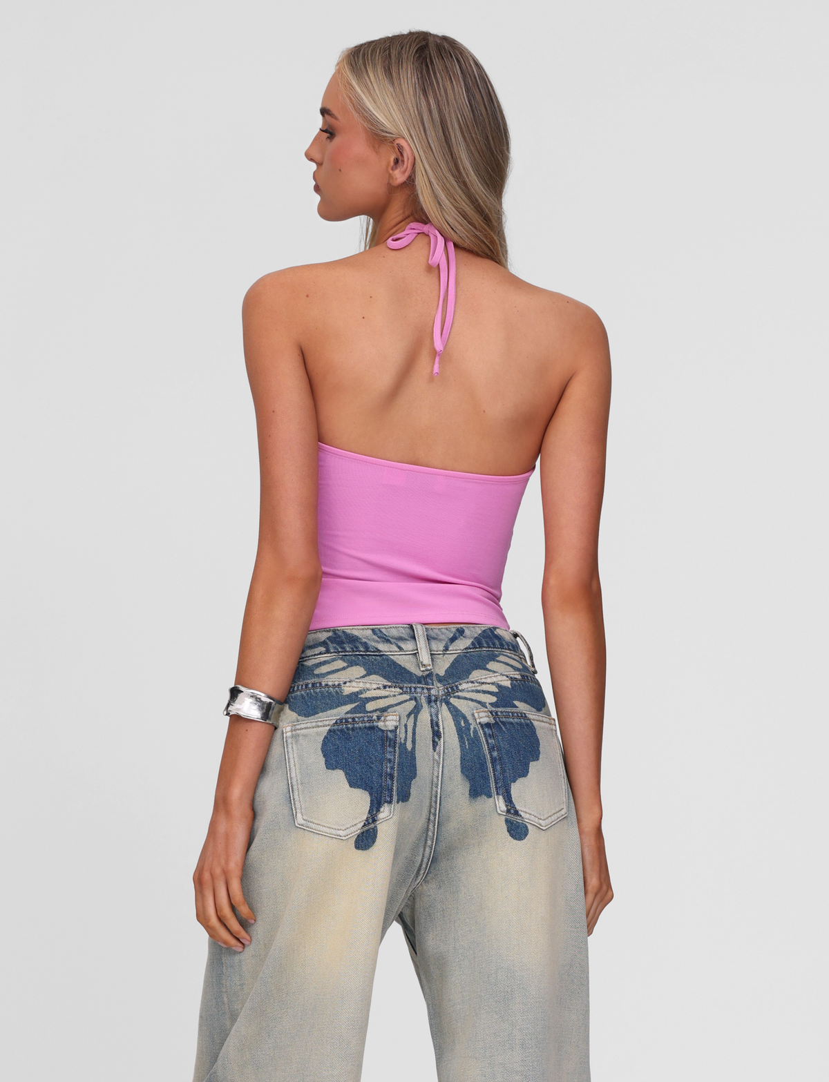 A woman with long blonde hair faces away from the camera, wearing the TIGERMIST CELESTINE TOP in pink and light blue baggy jeans featuring a large dark blue butterfly print on the back.