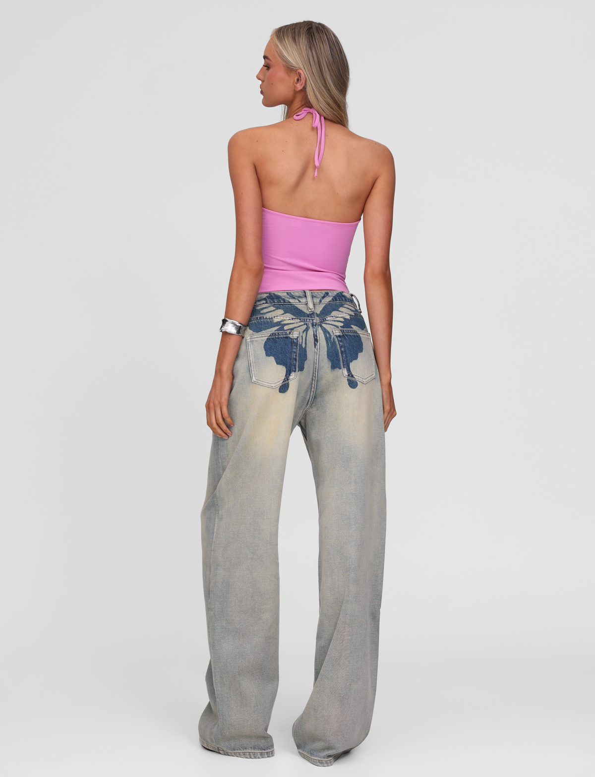 A woman with long hair wears TIGERMIST's ZANTHE JEAN - GREY WASH, featuring oversized, grey-wash denim and dark blue butterfly prints on the back pockets. She pairs it with a pink halter top and a silver bracelet, standing against a plain white background.
