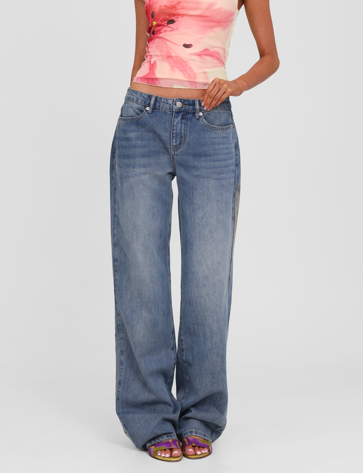 A person models TIGERMIST’s ESTRELLA JEAN - MID WASH, styled with a sleeveless floral top and purple open-toed sandals, standing before a white backdrop. The face is not shown.