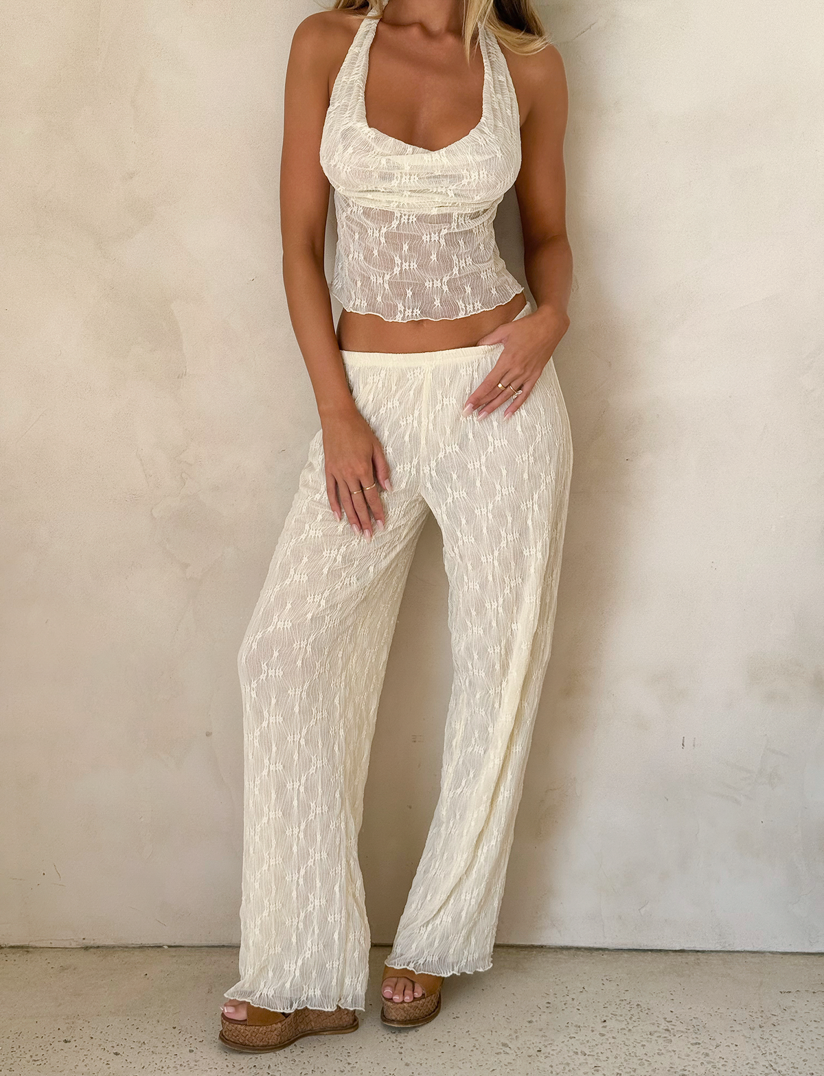 A woman, her hair down and face out of frame, poses against a beige wall in the TIGERMIST KATERINA PANT - IVORY LACE, styled with a sheer sleeveless halter top and tan platform sandals.