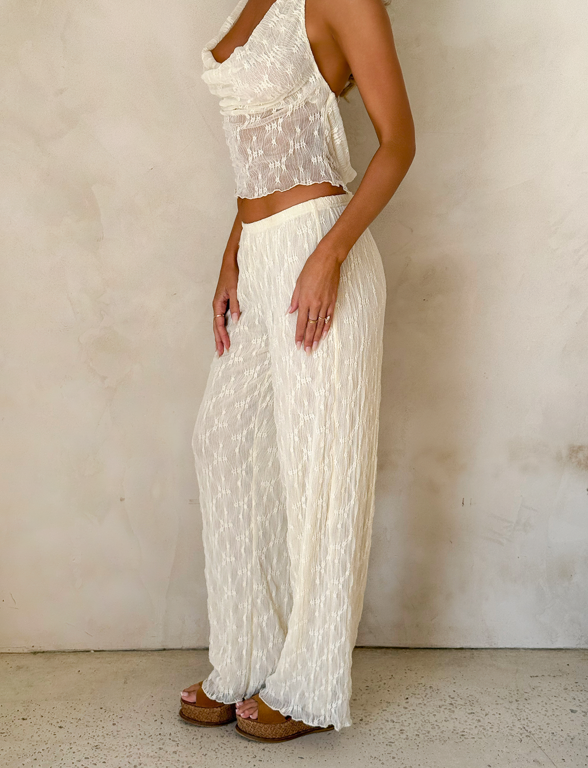 A woman stands against a light wall wearing TIGERMIST's KATERINA PANT in ivory lace with a sleeveless, semi-sheer cowl-neck top and brown platform sandals.