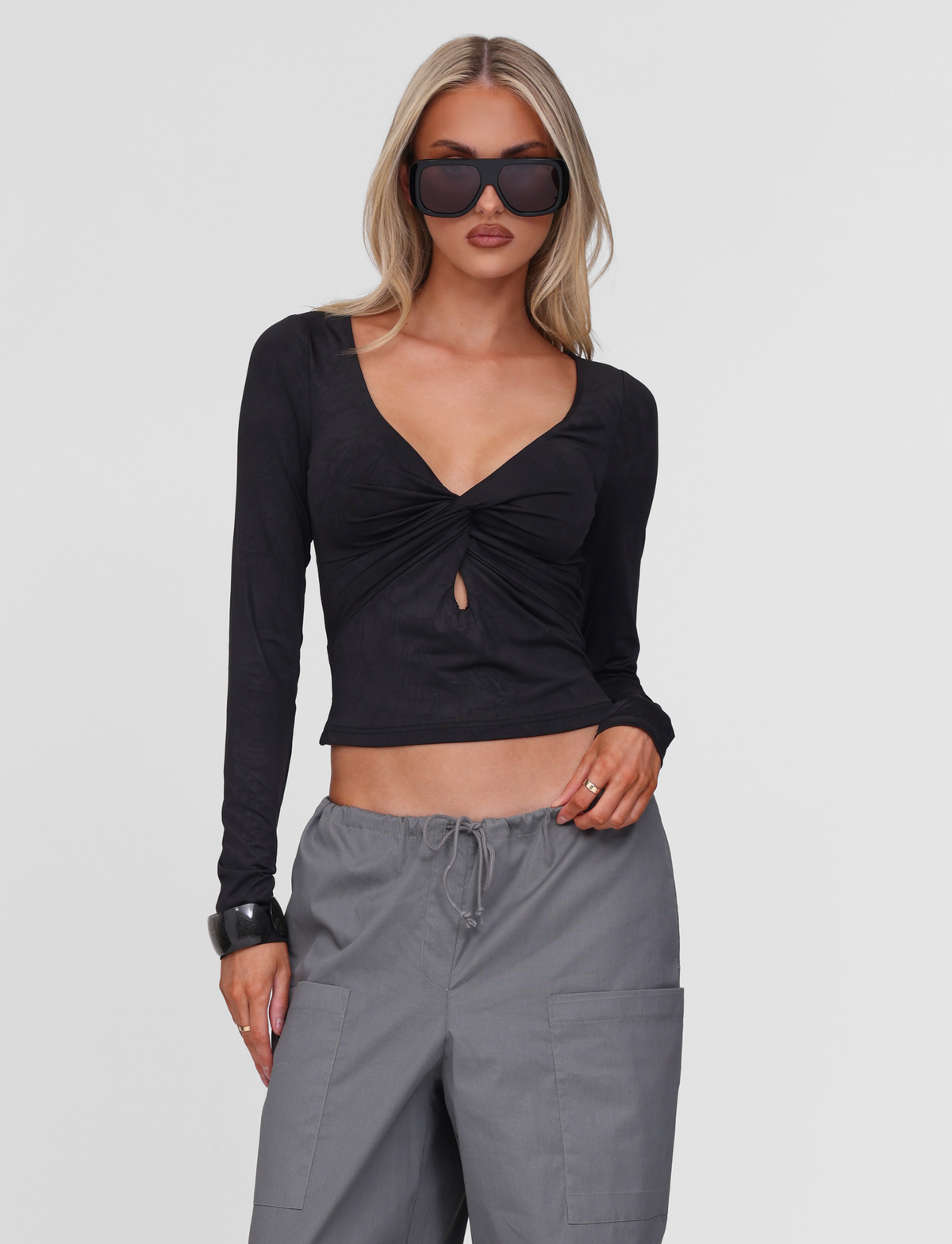 A woman wears the TIGERMIST SAFFIRA LONG SLEEVE TOP in black with oversized sunglasses and loose gray drawstring pants, standing against a plain white background.