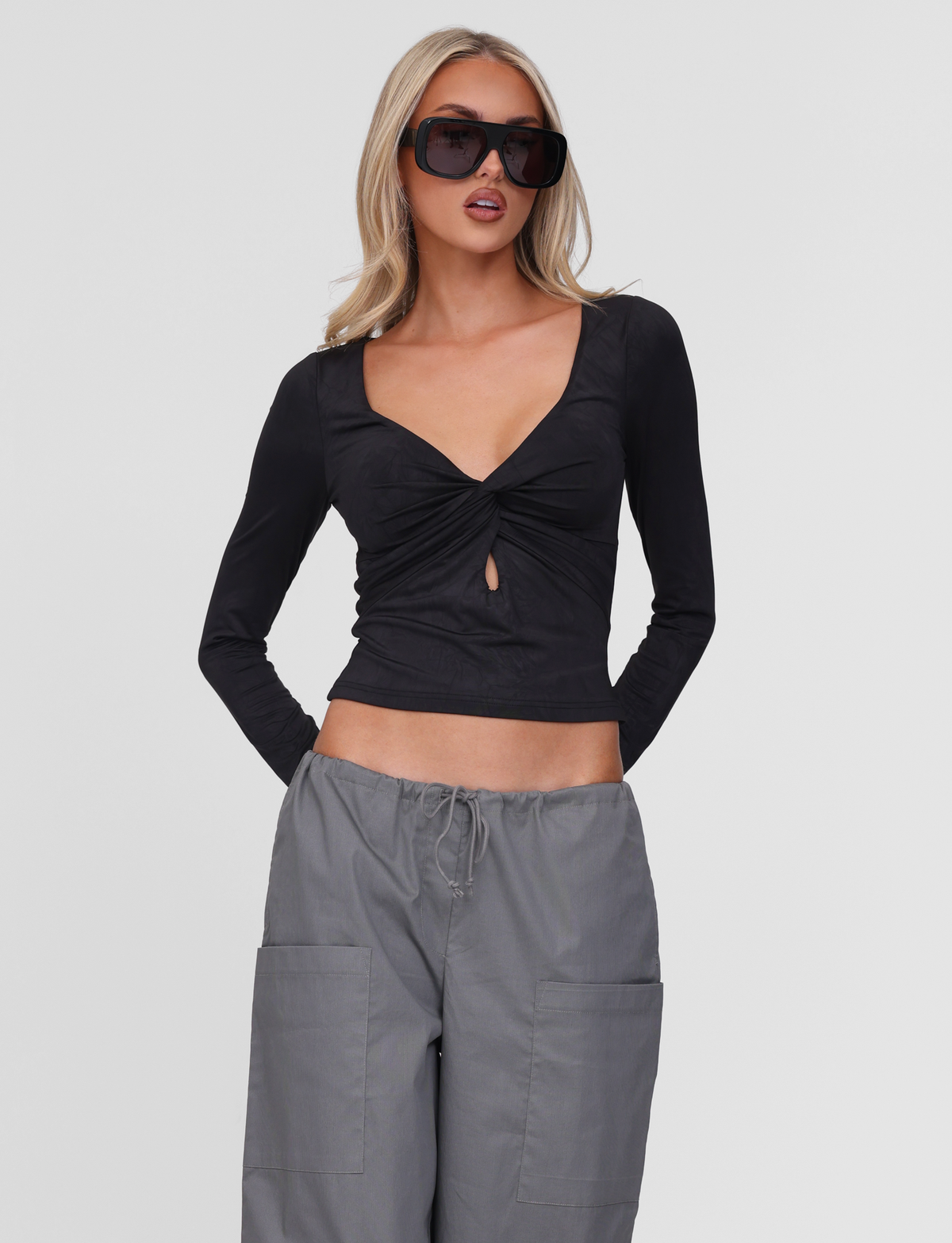 A woman wears the TIGERMIST SAFFIRA LONG SLEEVE TOP in black, featuring a twist front and keyhole V neckline, paired with loose gray pants and oversized sunglasses as she poses against a plain white background.