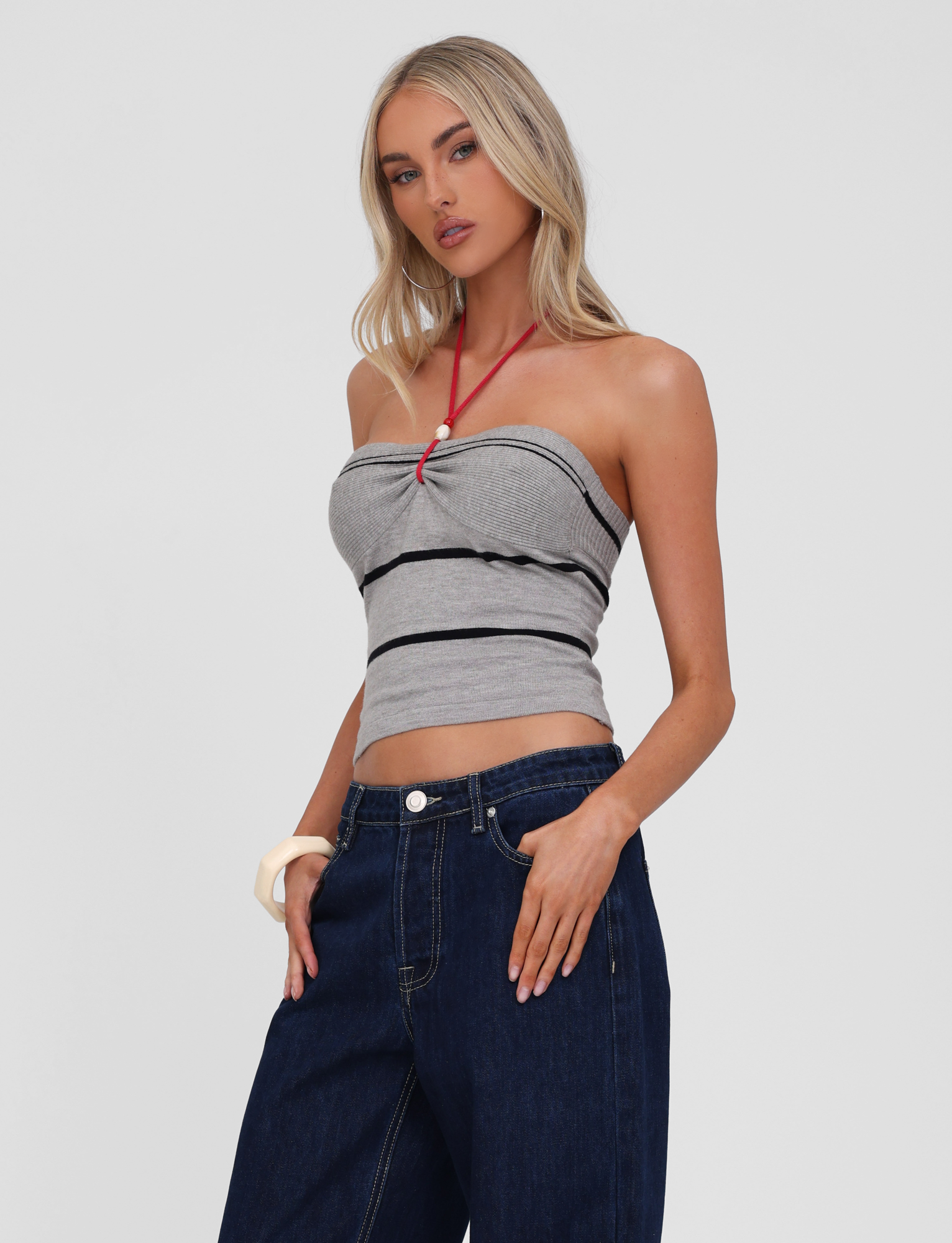 A woman with long blonde hair models the TIGERMIST NOUR TOP - GREY STRIPE, a stripy tube top, paired with wide-leg dark blue jeans. She poses with one hand in her pocket against a plain light background.