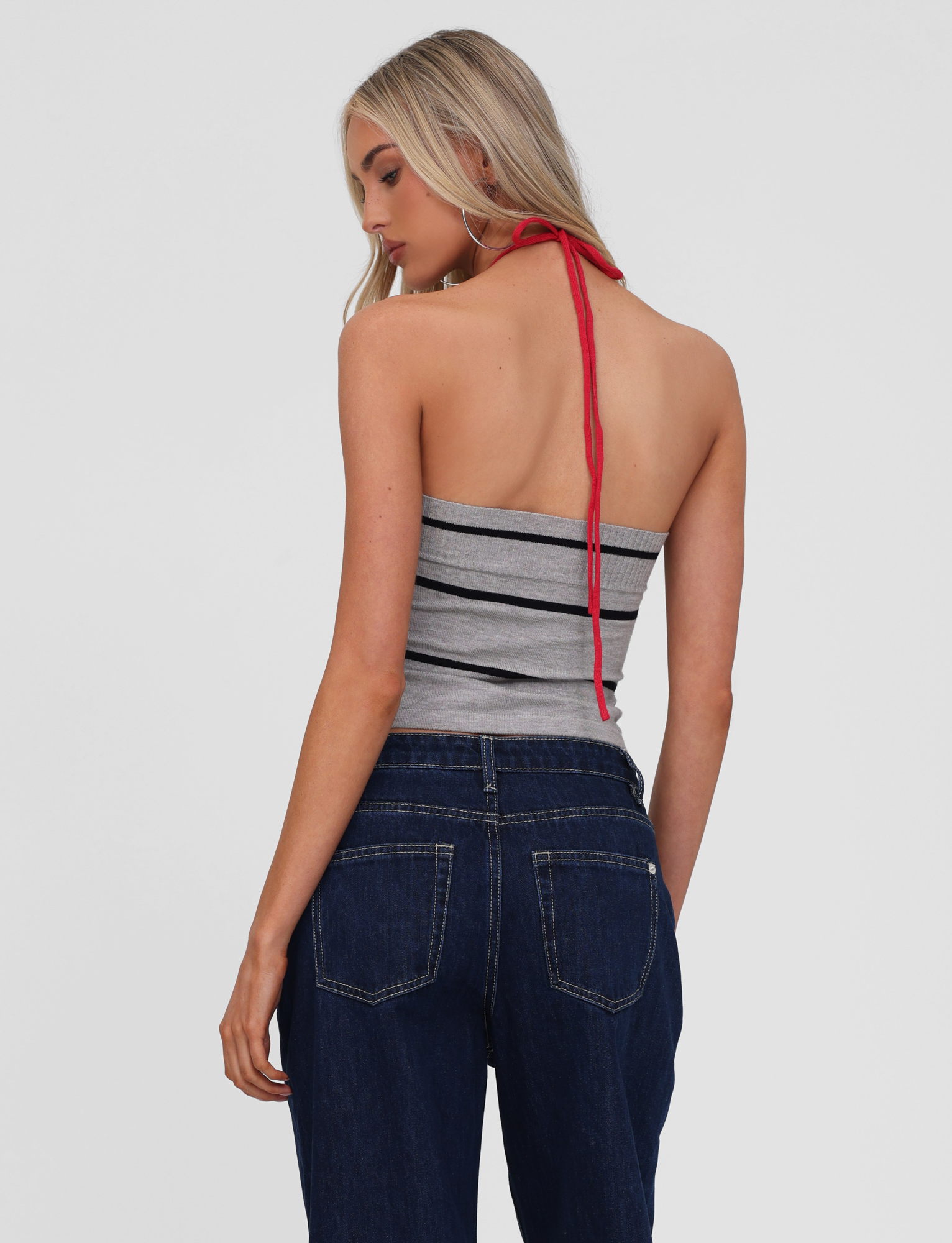A woman with long blonde hair is seen from the back, wearing the TIGERMIST NOUR TOP - GREY STRIPE with red halter ties and dark blue jeans, standing against a plain light background.
