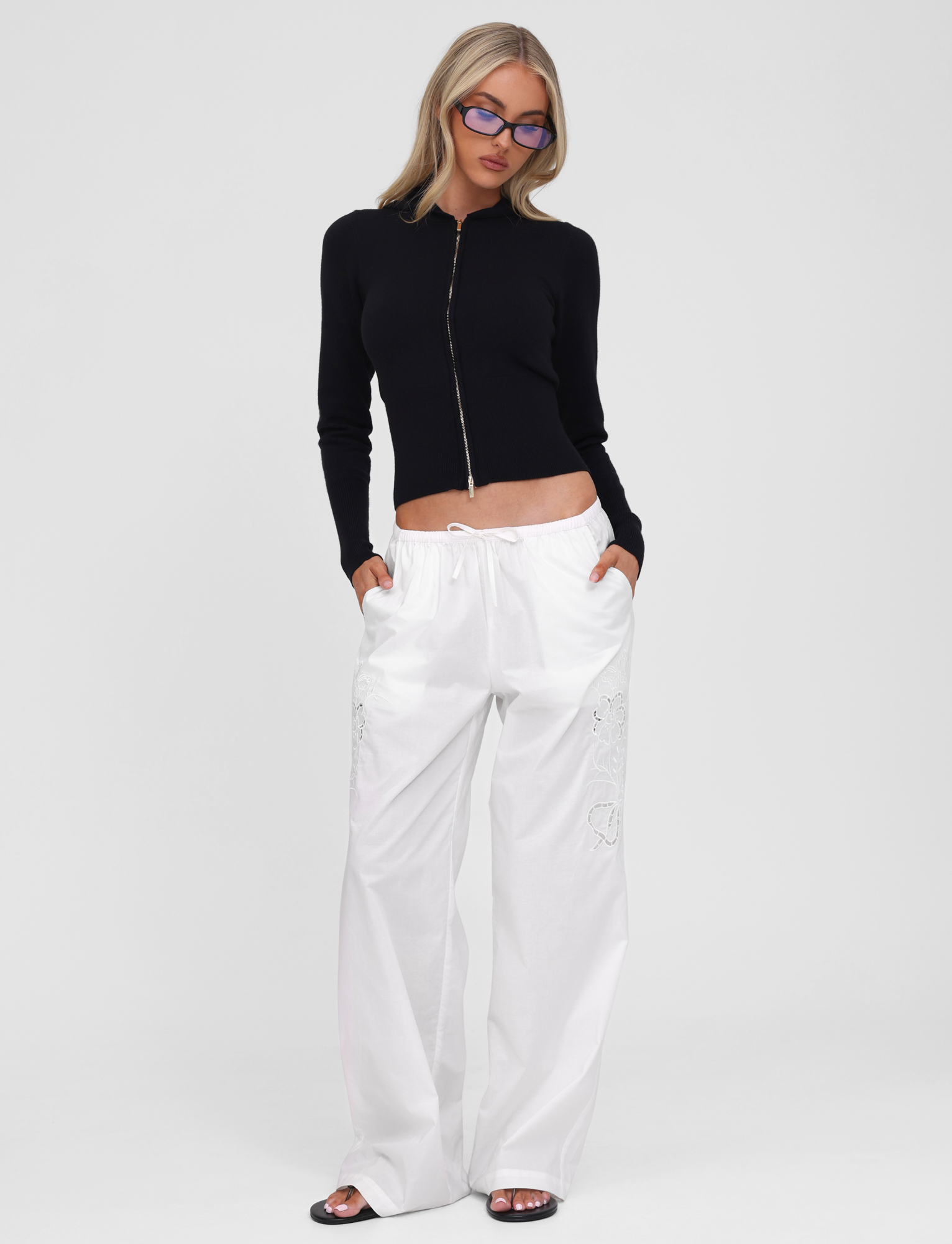 Wearing the TIGERMIST SARINA JUMPER in black, paired with loose white pants, black slides, and blue-tinted glasses, a person stands against a plain white background.