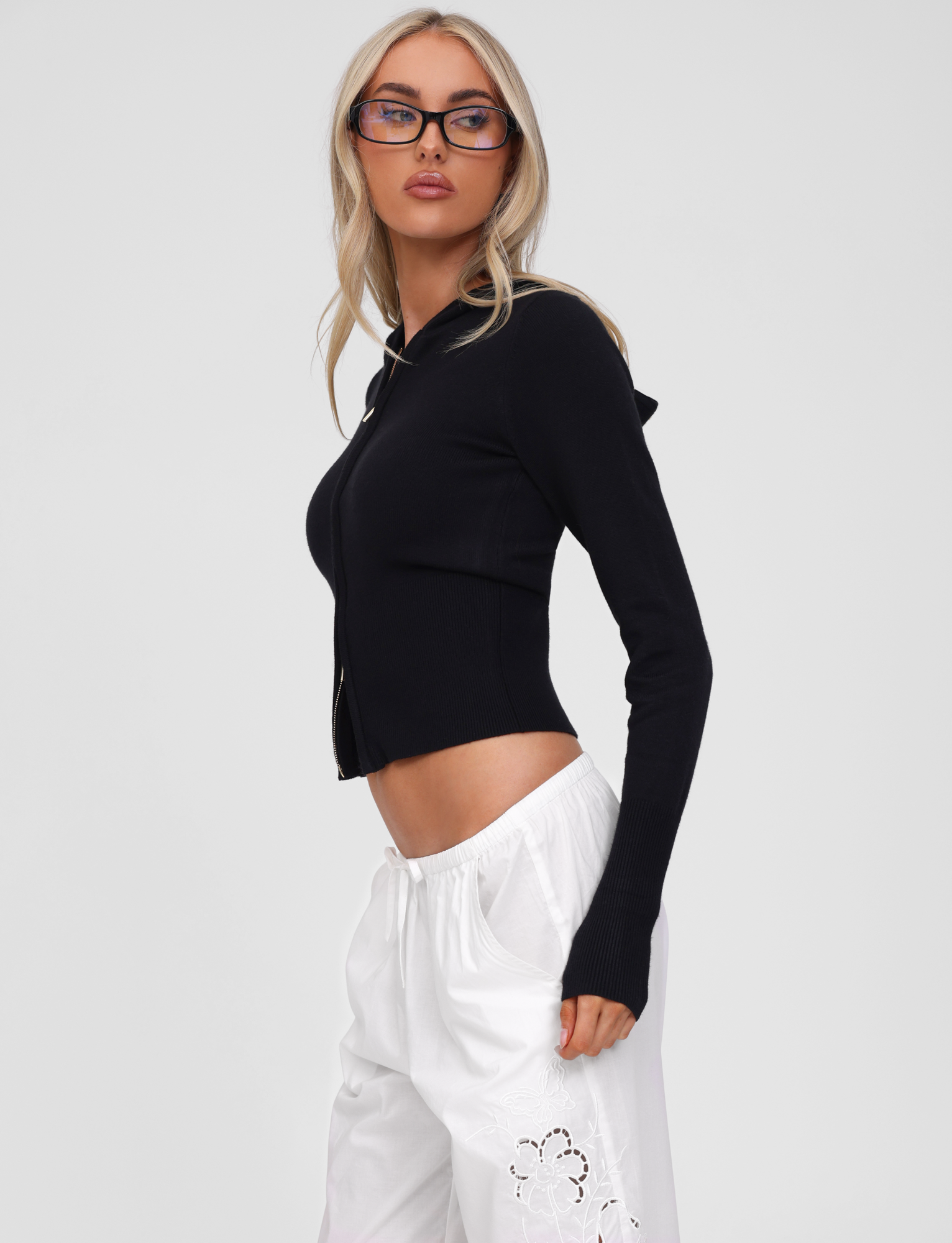 A woman with long blonde hair and glasses poses against a plain light background wearing the TIGERMIST SARINA JUMPER - BLACK, a fitted black knit hoodie with a gold zipper, paired with loose white pants with floral cutout details.