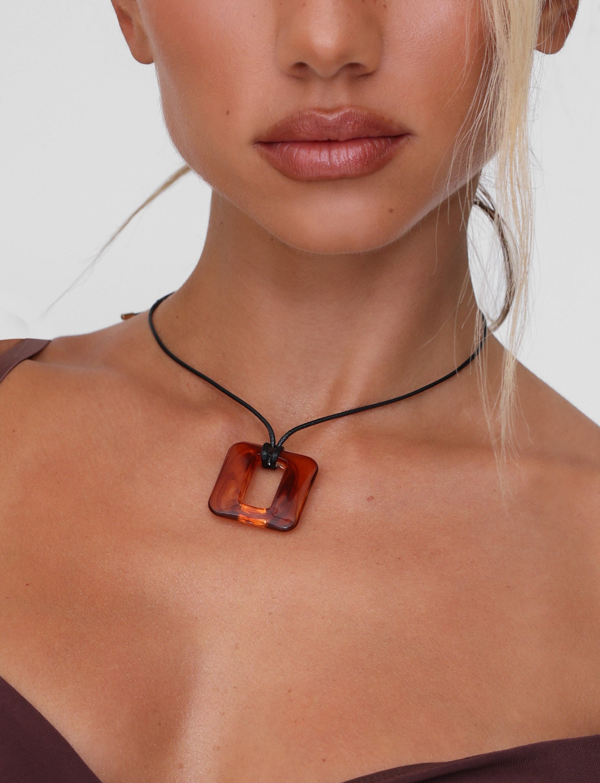 A person wearing the TIGERMIST MIRREN SQUARE NECKLACE in brown, featuring an amber pendant, adjusts the cord to complement their long hair and brown top with thin straps against a plain white background.