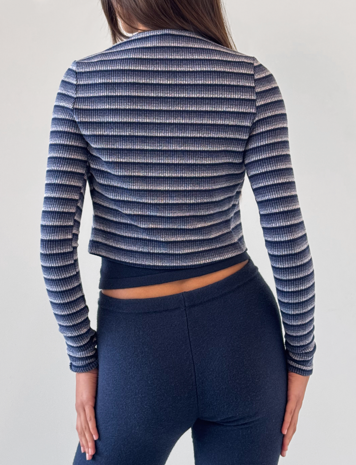 A woman with straight brown hair faces away, wearing TIGERMIST's LEORA PANT in navy, paired with a navy and white striped long-sleeve top. The pants feature a relaxed fit and drawstring waist. The background is plain and light-colored.