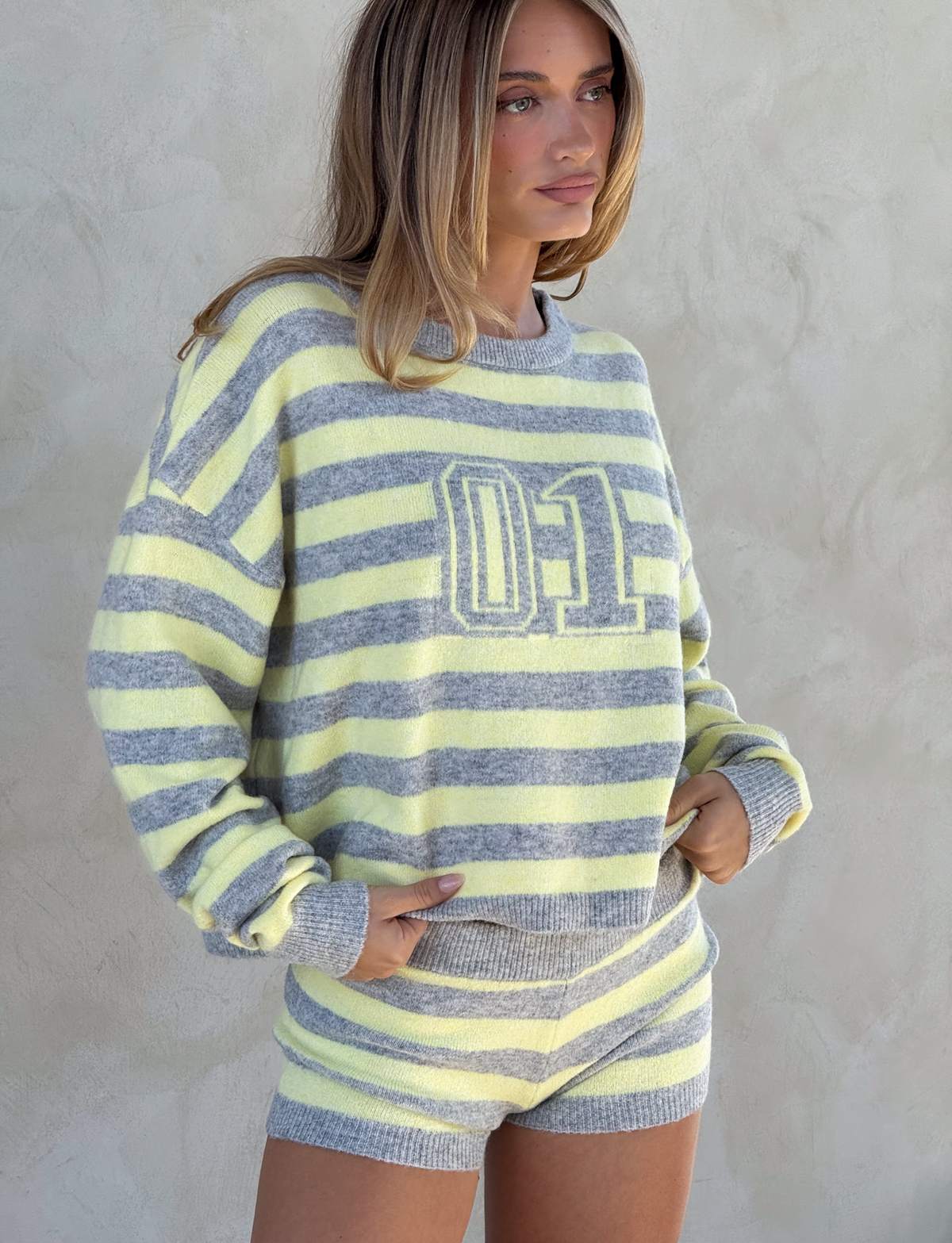A woman poses against a light textured wall in the TIGERMIST KOA JUMPER, an oversized knit featuring yellow and grey stripes with a sporty "01" print, paired with matching shorts and her hands in the pockets.