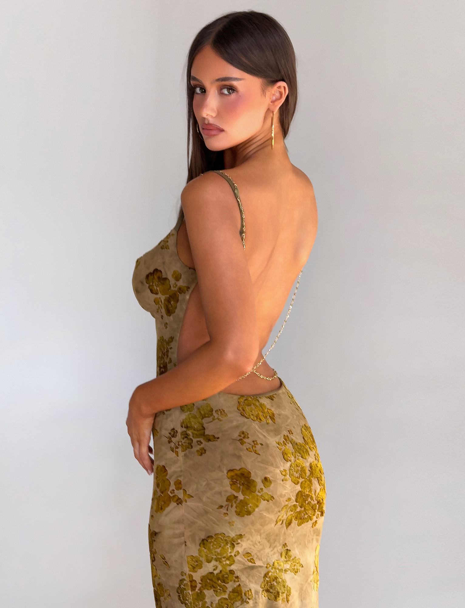 A woman with long brown hair wears the TIGERMIST SPARROW MAXI DRESS - GREEN BURNOUT, featuring a green and gold floral print. The elegant backless design stands out as she poses sideways against a neutral background.