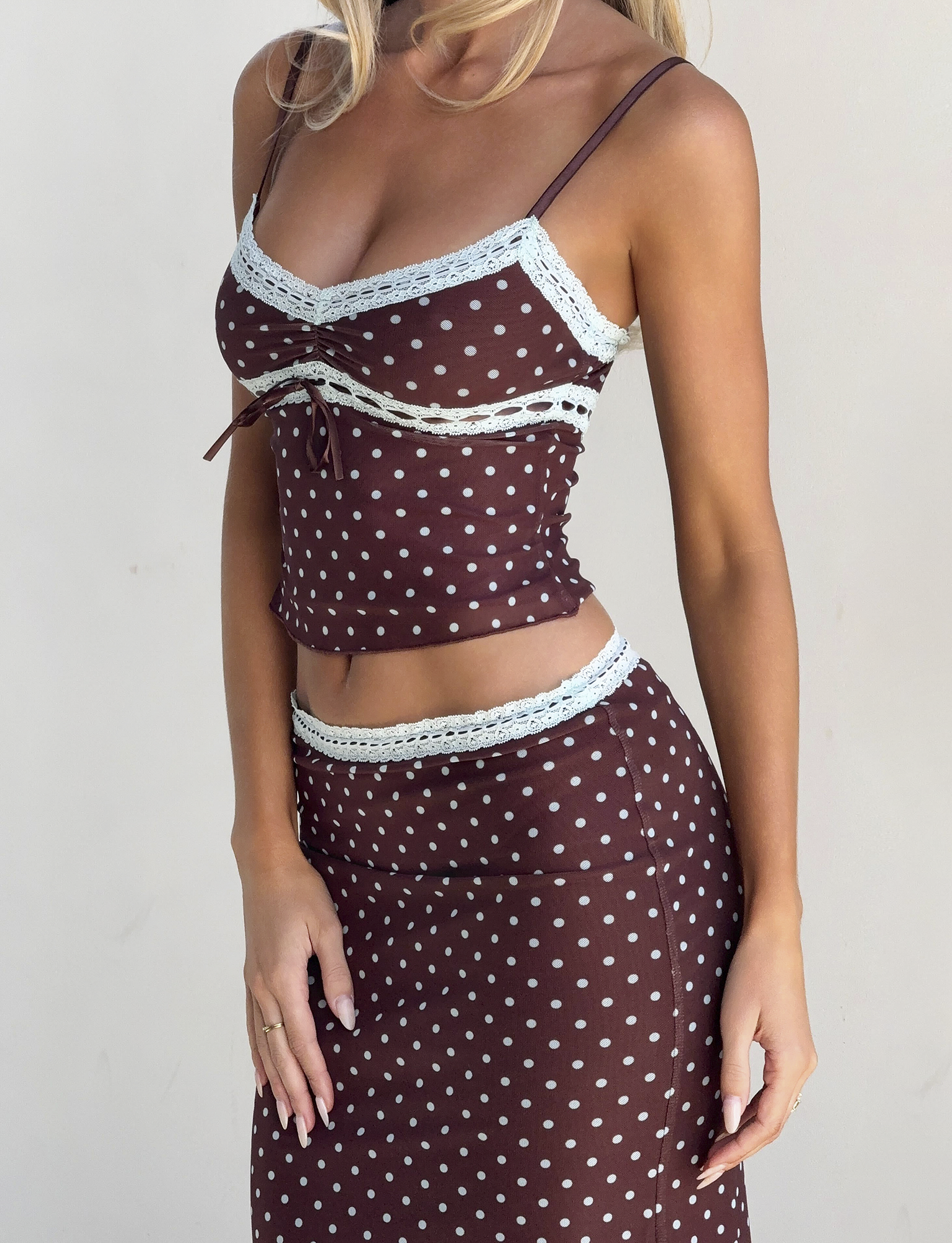 A woman poses against a plain light background, wearing the TIGERMIST AMARA MIDI SKIRT - POLKA DOT, paired with a brown camisole top and delicate lace details.