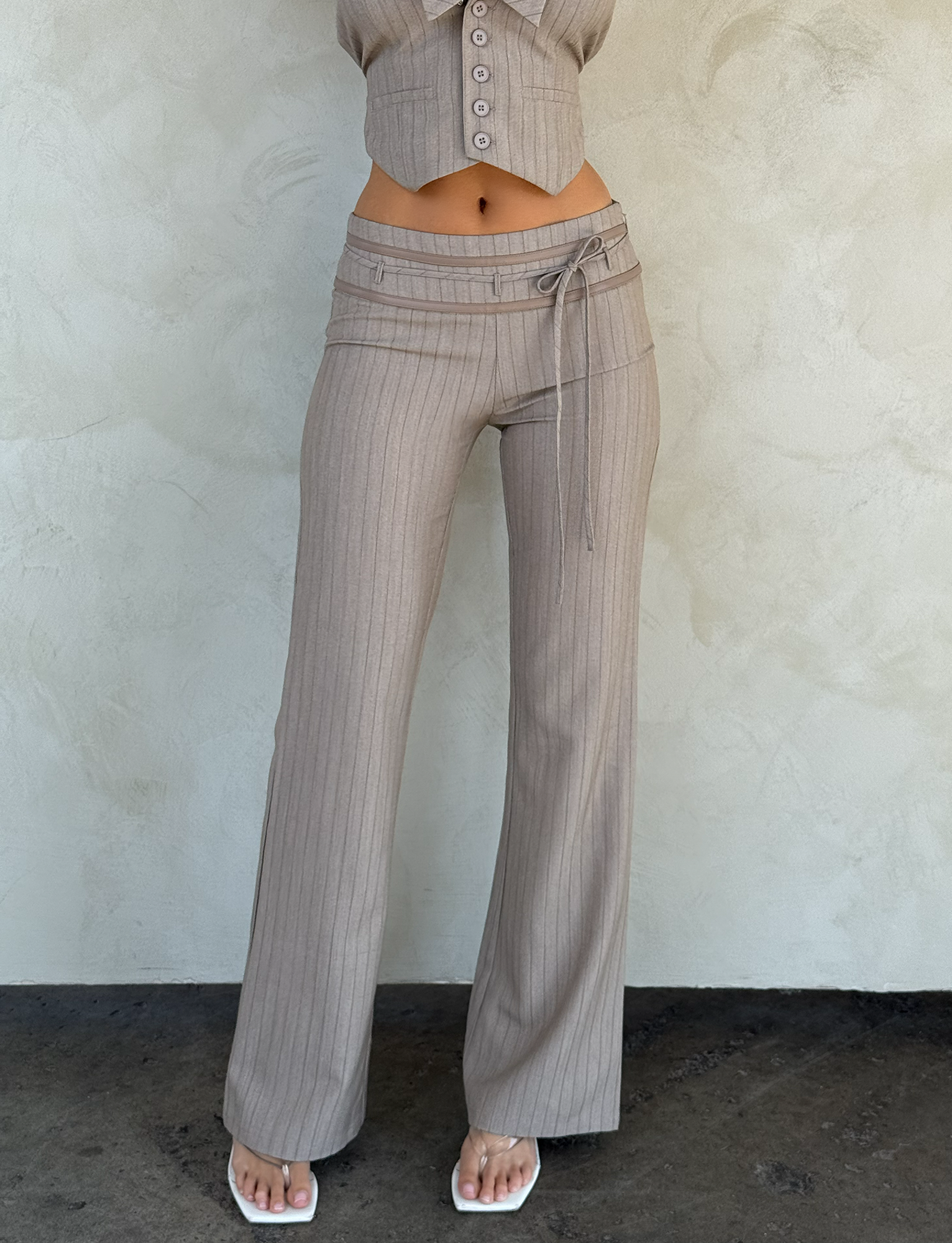 Wearing TIGERMIST's FEDRA PANT in Tan Pinstripe with a matching cropped top and white square-toe sandals, this person poses against a textured light-colored wall.