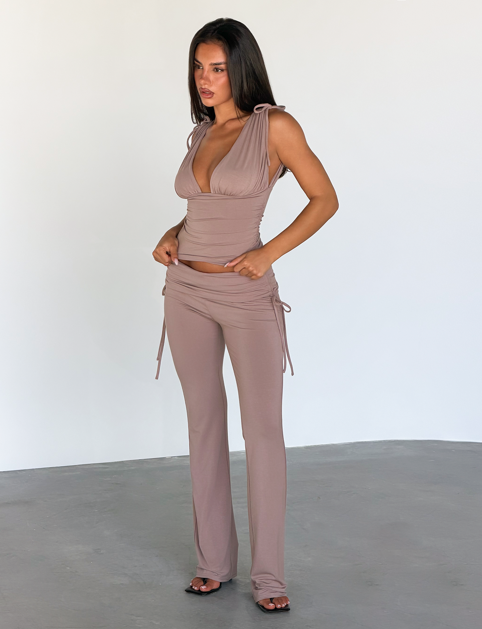A woman stands indoors wearing the TIGERMIST HALONA PANT in mocha—a flared pant—paired with a sleeveless, taupe V-neck top. She faces slightly left, arms relaxed at her sides.