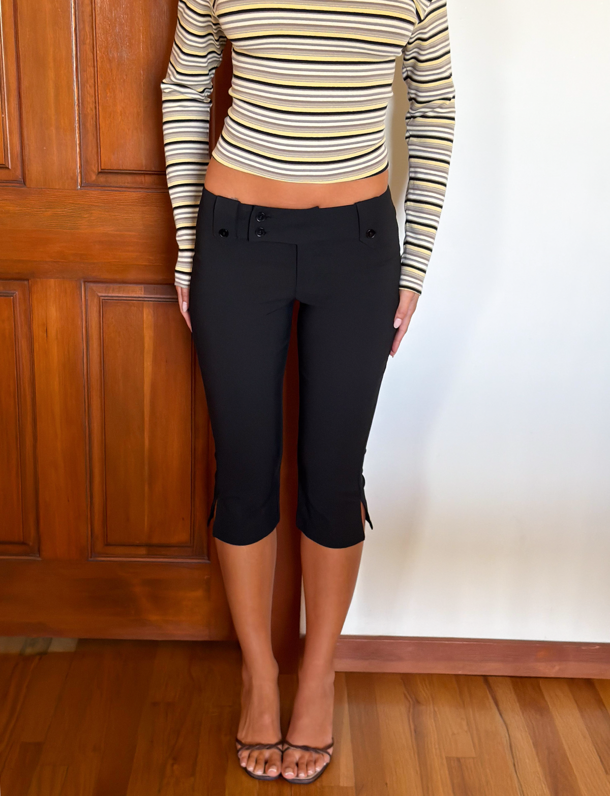 A person (shoulders down visible) stands indoors by a wooden door and white wall, wearing TIGERMIST’s KITTIE CAPRI - BLACK featuring a wide crossover waistband and button details, paired with a long-sleeve striped crop top.