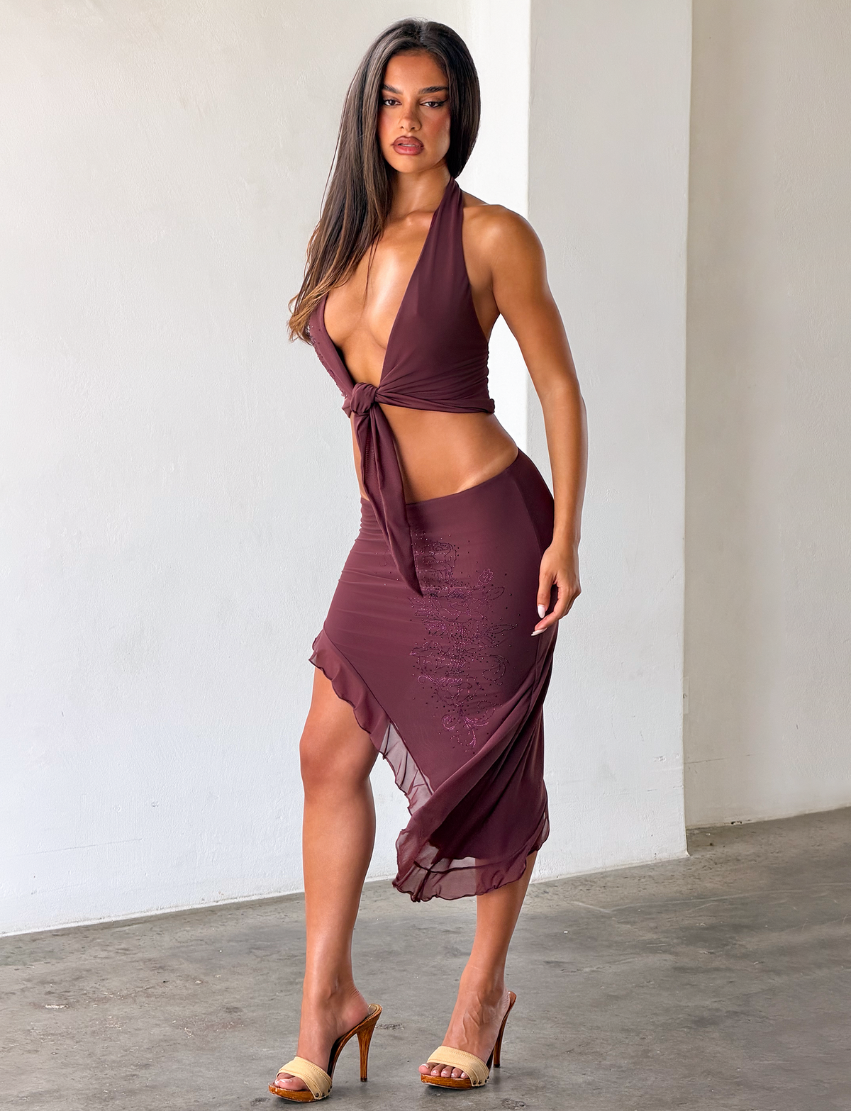 A woman with long brown hair poses confidently indoors in tan high-heeled sandals, pairing the TIGERMIST GEORGIE DIAMANTE HI LO SKIRT in Chocolate with a sleeveless halter top.