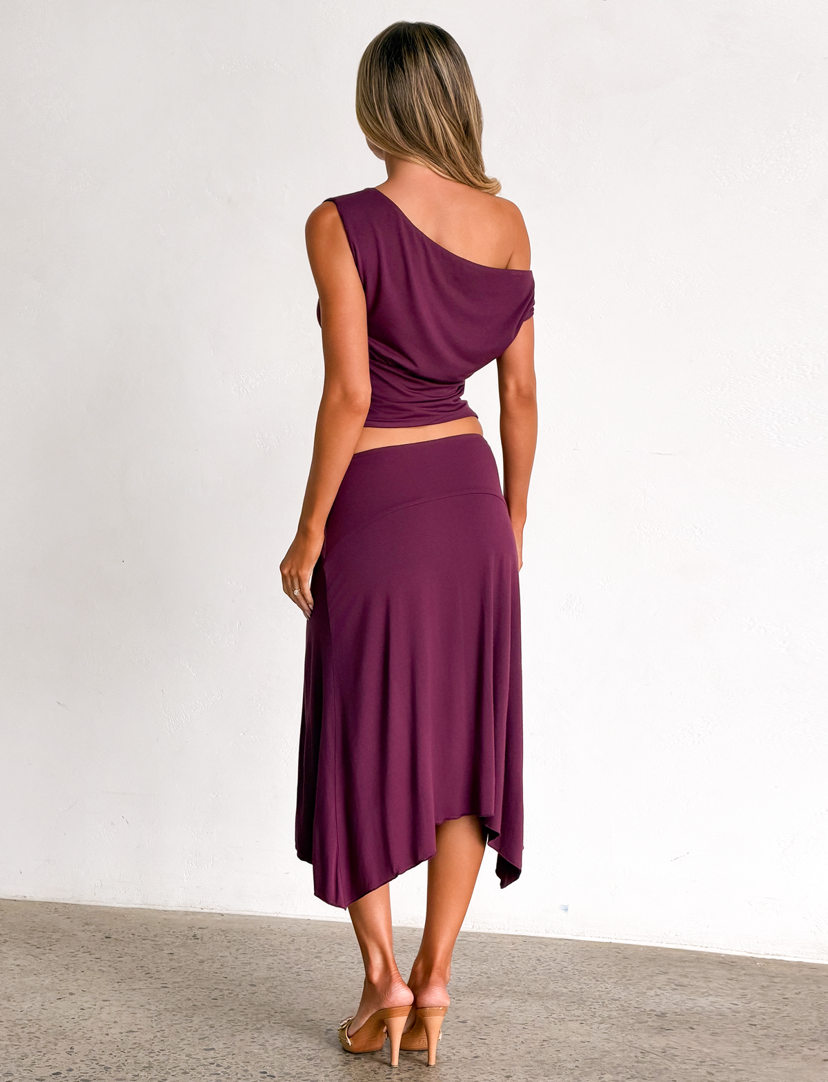 A woman is seen from behind wearing a purple one-shoulder top with the TIGERMIST ALYSSA HI LO SKIRT in plum, featuring an asymmetrical hem.