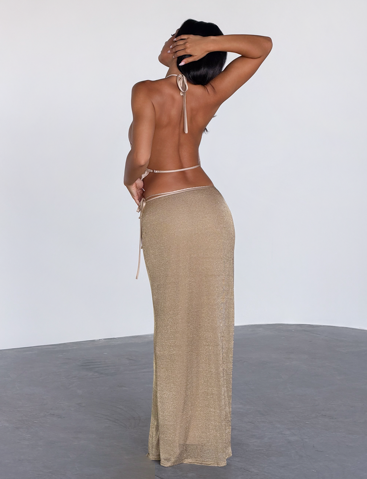 A woman stands on a gray floor facing away, wearing the TIGERMIST CYRENE SATIN COWL TOP in gold with halter straps and a matching long skirt. One arm is raised to her head, the other rests on her hip. The background is plain and light-colored.