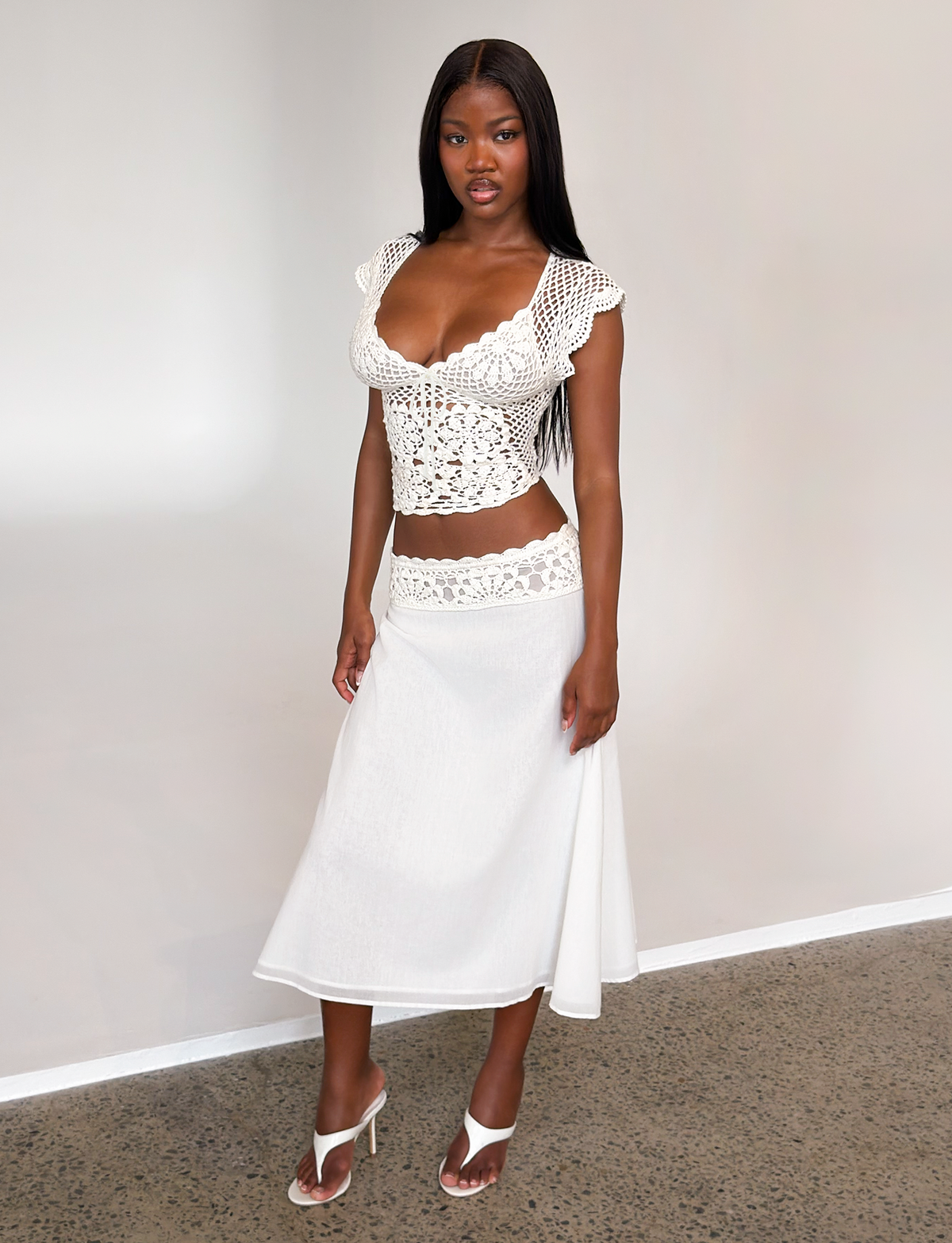 A woman stands indoors against a light background wearing the TIGERMIST SASHA CROCHET MIDI SKIRT in ivory, styled with a white crocheted cap-sleeve top and white heeled sandals.