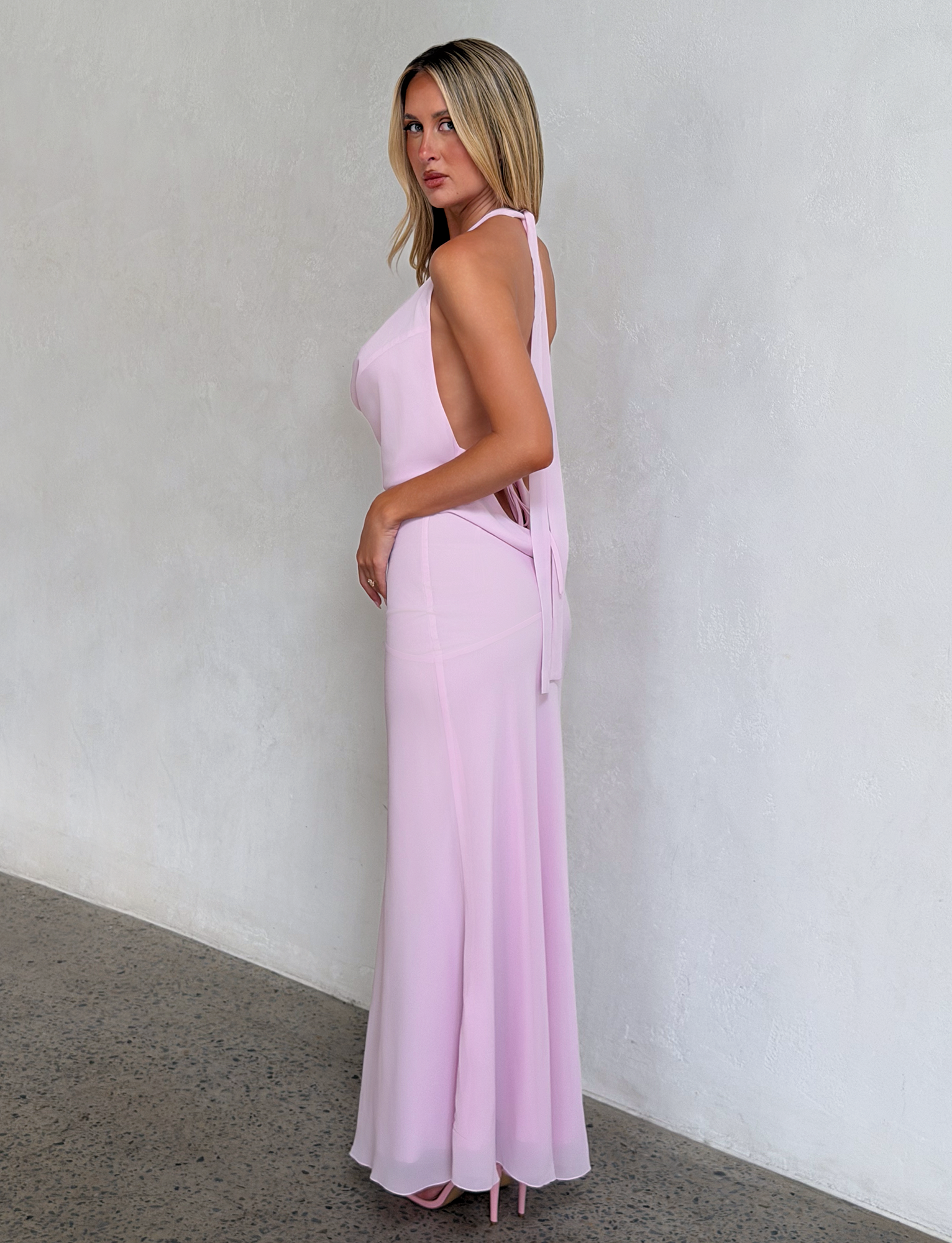A woman with straight blonde hair wears the TIGERMIST ZYLA MAXI DRESS in baby pink and matching heels, standing sideways against a light-colored textured wall while looking at the camera.