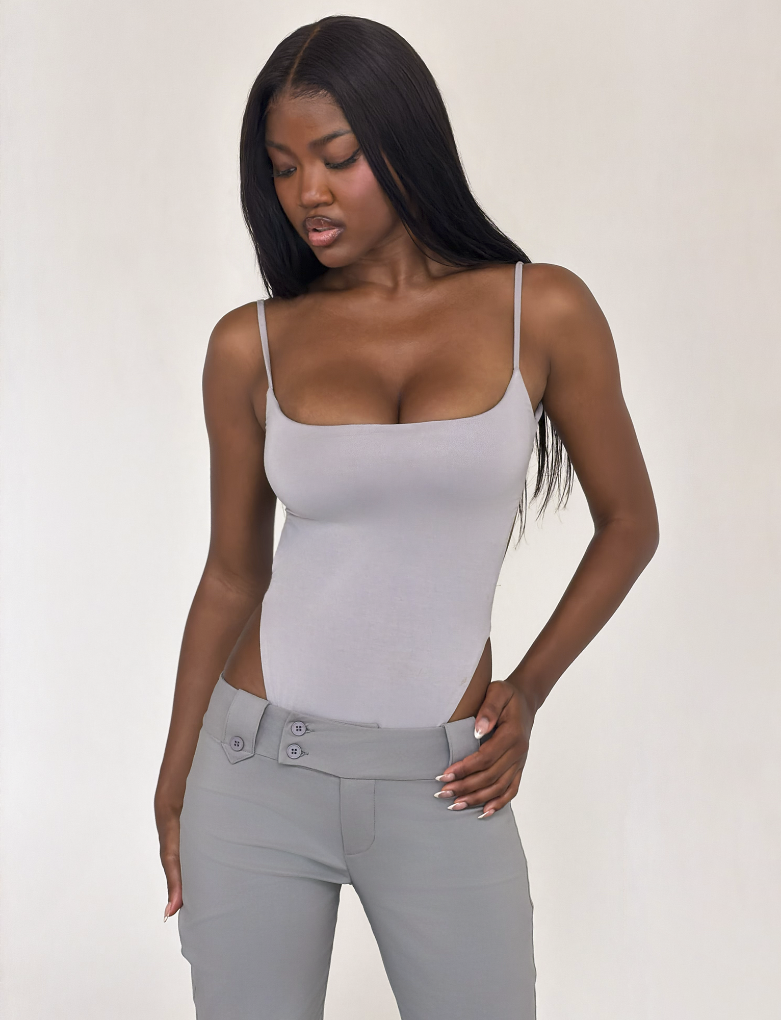 A woman with long straight black hair wears the TIGERMIST MINERVA BODYSUIT in Frost Grey and low-rise gray pants, standing against a plain light background while looking down to her right.