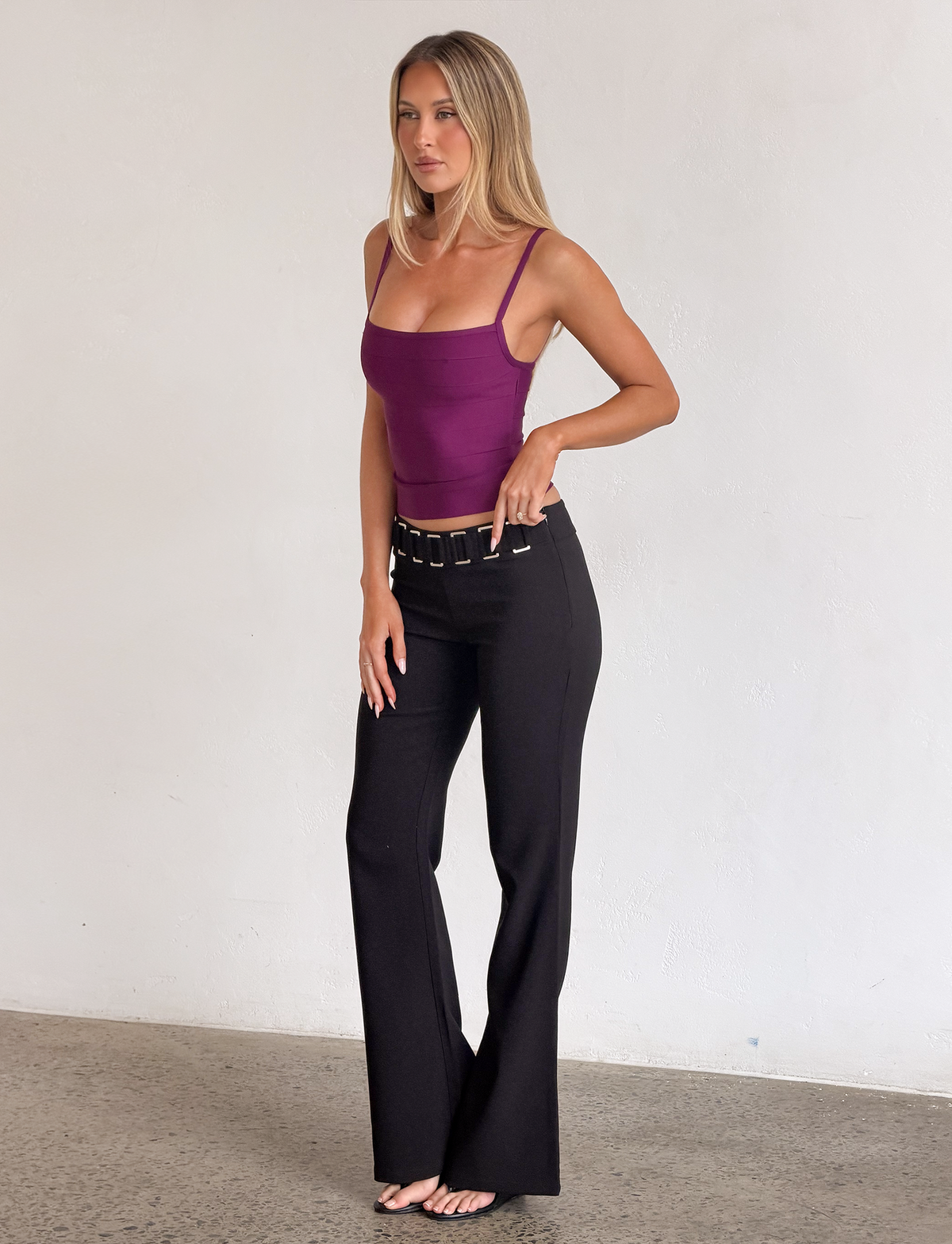 A woman stands indoors posing against a plain white wall on a concrete floor, wearing the TIGERMIST MIDORI BANDAGE TOP in plum—a bandage tank top with a square neckline—paired with high-waisted black pants and a wide belt.