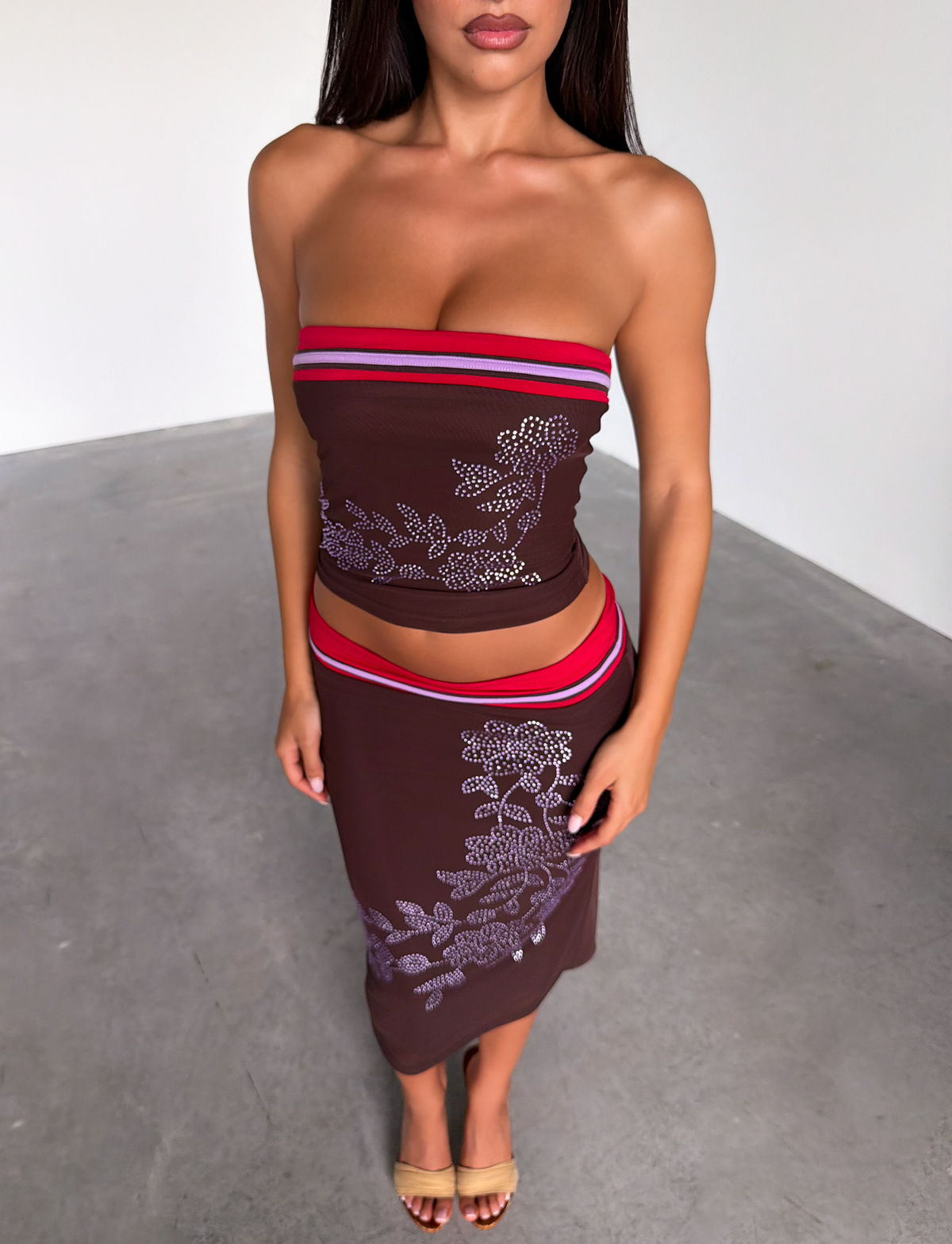 A woman stands indoors on a concrete floor wearing the TIGERMIST SERAH DIAMANTE BOOB TUBE in chocolate with a matching skirt, featuring red and white floral patterns; her head is partially out of frame.