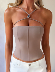 A woman wears the TIGERMIST TAMORA CORSET TOP in taupe, featuring a 90’s-inspired design with crossover straps and a blue center pendant, paired with high-waisted white pants. Her face is not visible.