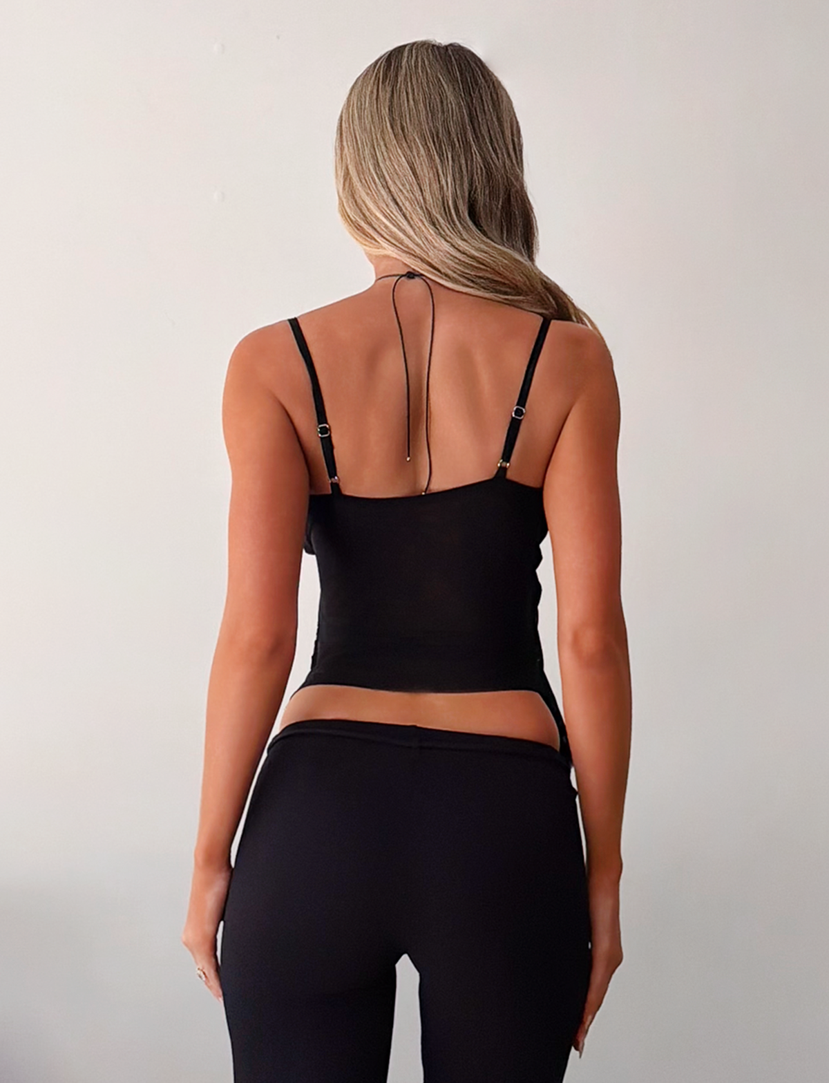 A woman with long, wavy blonde hair stands with her back to the camera, wearing the TIGERMIST ALDEANA TOP in black and black pants against a plain, light background.