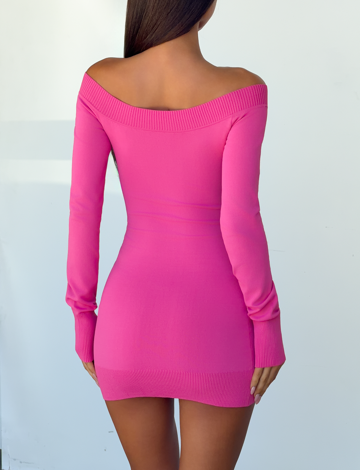 A woman stands facing away, wearing the PARKER KNIT MINI DRESS in fuchsia by TIGERMIST. The fitted, long sleeve knit dress features an off-the-shoulder design. Her straight brown hair falls down her back against a plain white background.