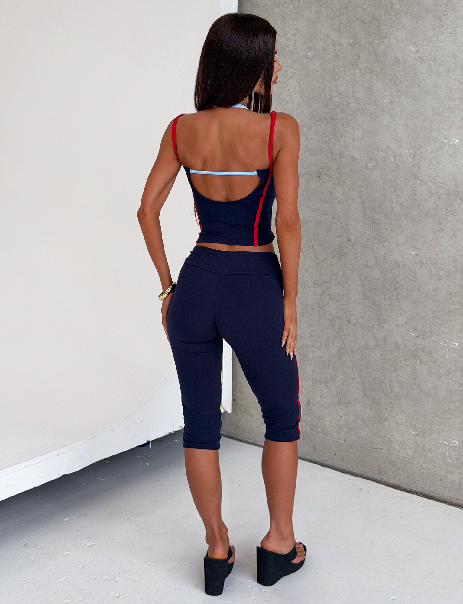 A woman with long brown hair stands indoors facing away, wearing TIGERMIST's TATUM CAPRI - NAVY STRIPE pants and a matching crop top with red accents, paired with black platform sandals against a plain gray textured wall.