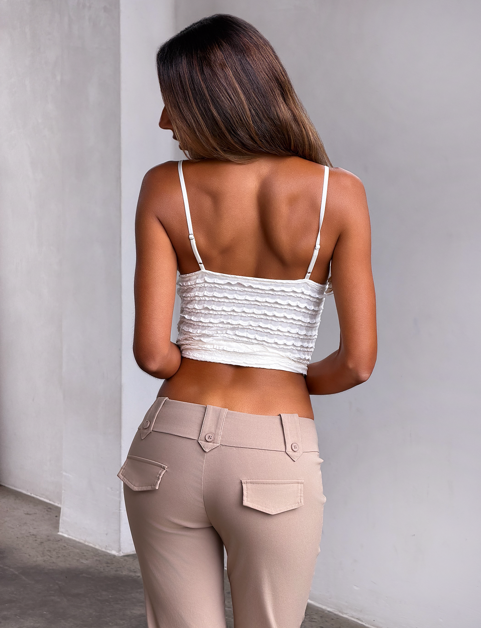 A woman with long brown hair, wearing the TIGERMIST ZORA TOP in cream with adjustable straps and beige pants, stands with her back to the camera against a light-colored wall.