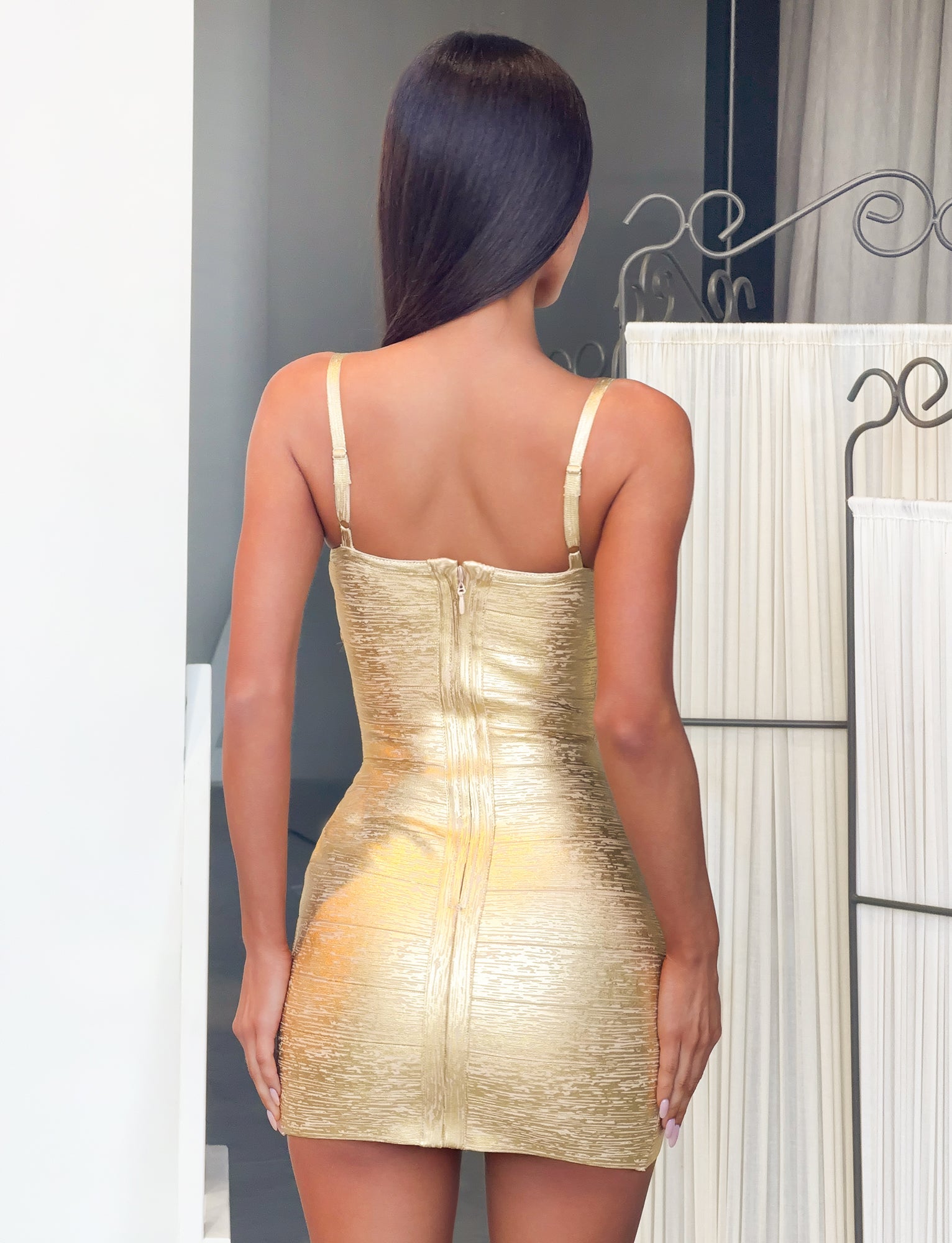 A woman with long dark hair stands facing away, wearing the TIGERMIST KENZA BANDAGE MINI DRESS in gold. This metallic, form-fitting mini dress features a bodycon silhouette, a zipper down the back, and a sleeveless design—making it perfect for special occasions. The setting includes neutral-toned walls and a decorative metal divider in the background.