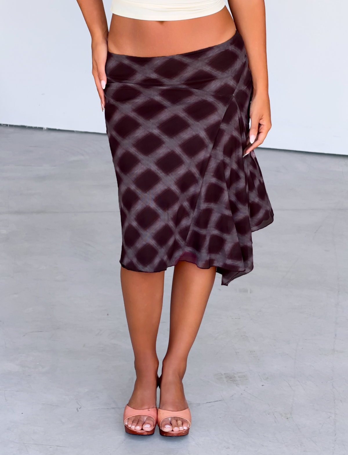 A person stands on a smooth, light gray floor wearing the TIGERMIST SAFFRON MIDI SKIRT in mahogany check and light pink open-toed heels. The upper body and face are not visible.