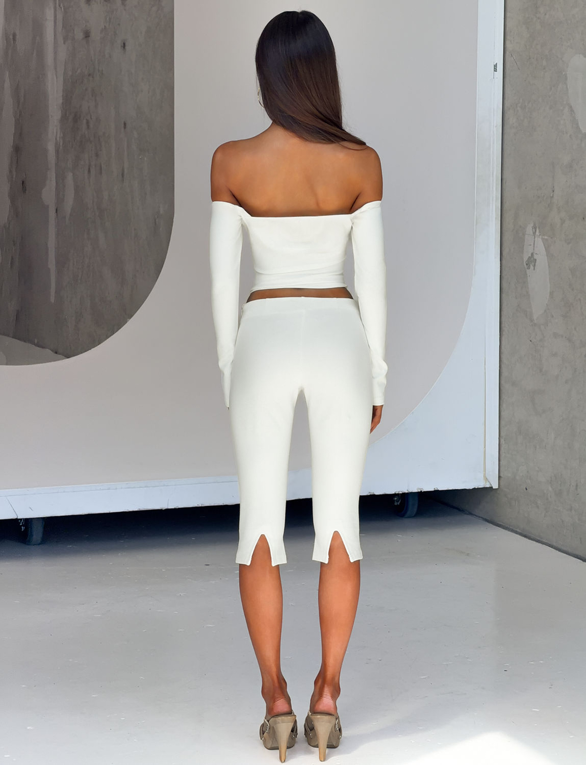 A woman stands indoors with her back to the camera, dressed in the TIGERMIST IDINA TOP - IVORY, an off-the-shoulder piece paired with matching high-waisted calf-length pants featuring small slits at the hems. Her long straight hair flows down her back and she finishes her sophisticated look with heeled sandals. Subtle faux pearl buttons on her ensemble add an elegant touch.