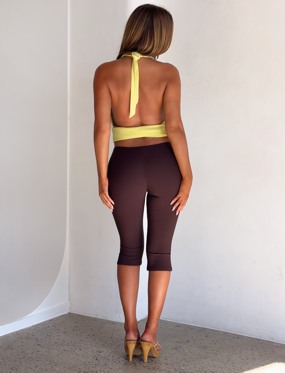 A person seen from behind wearing a TIGERMIST LYDIAH TOP - PISTACHIO halter top, dark brown capri leggings, and tan heeled mules.