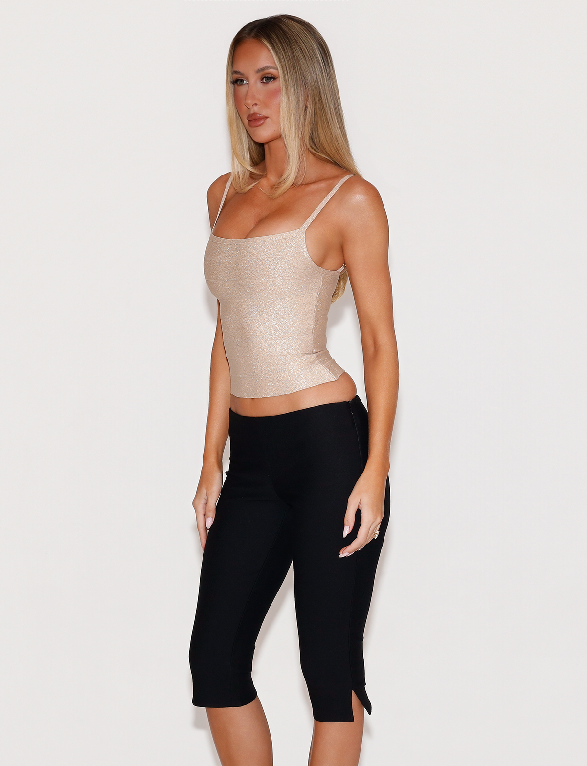 A woman wears a sparkly gold MIDORI BANDAGE TOP with a square neckline and thin straps, paired with black capri leggings.