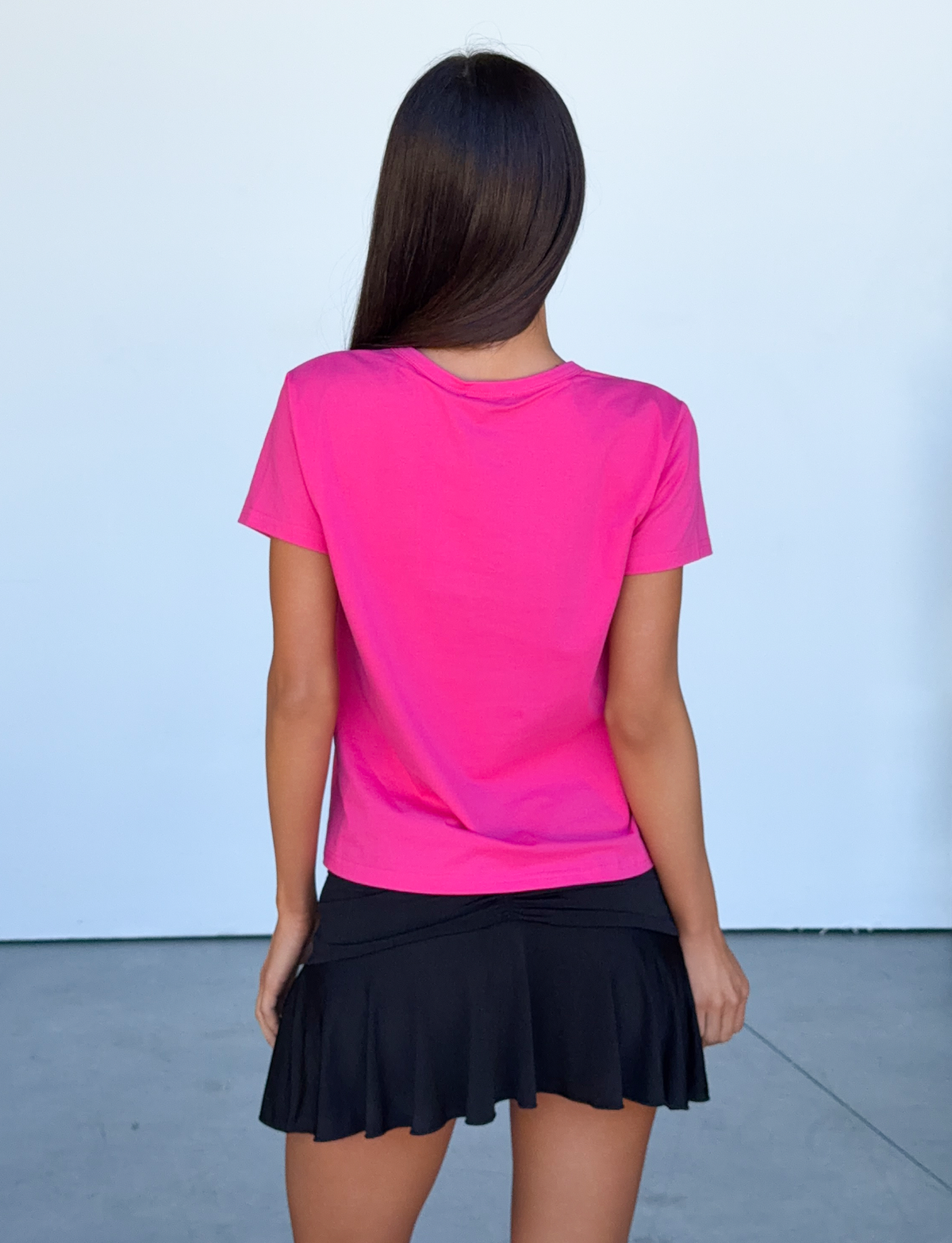 A woman with long brown hair stands facing away, wearing the TIGERMIST BELLA BABY TEE in hot pink and a short black skirt, against a plain light background.