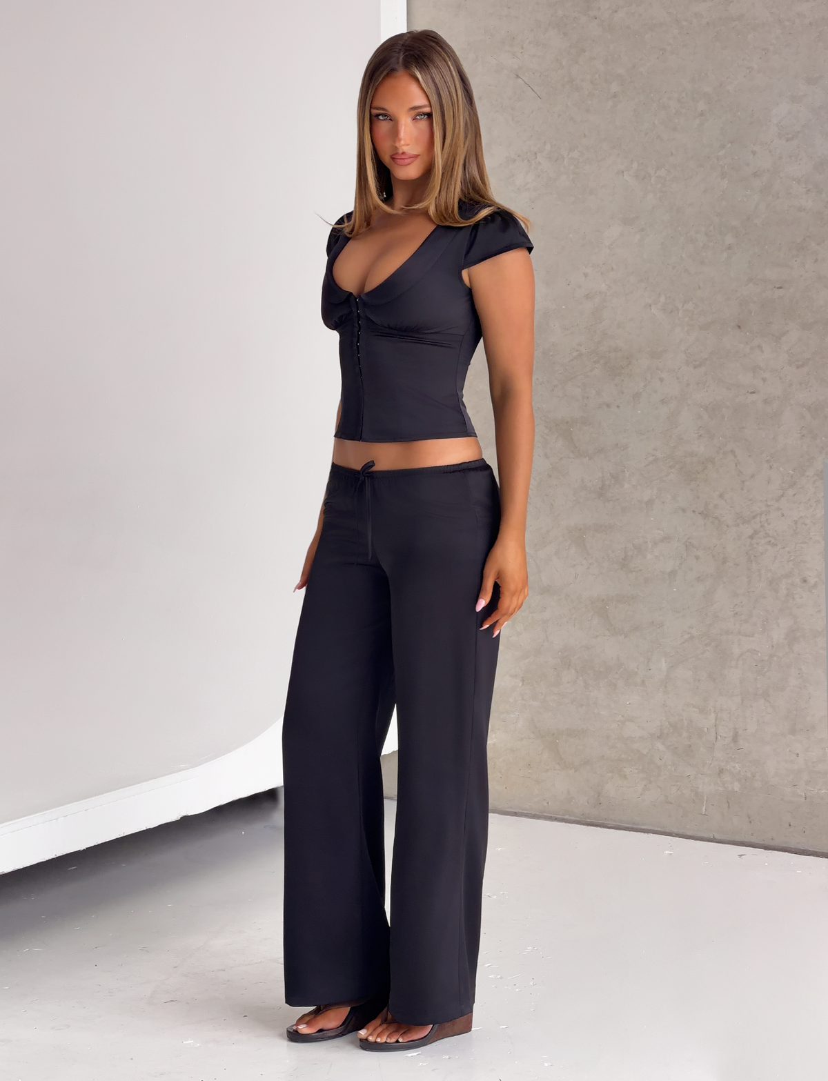 A woman wearing a black short-sleeved top with front-button detail and matching MAURA PANT - BLACK wide-leg pants.