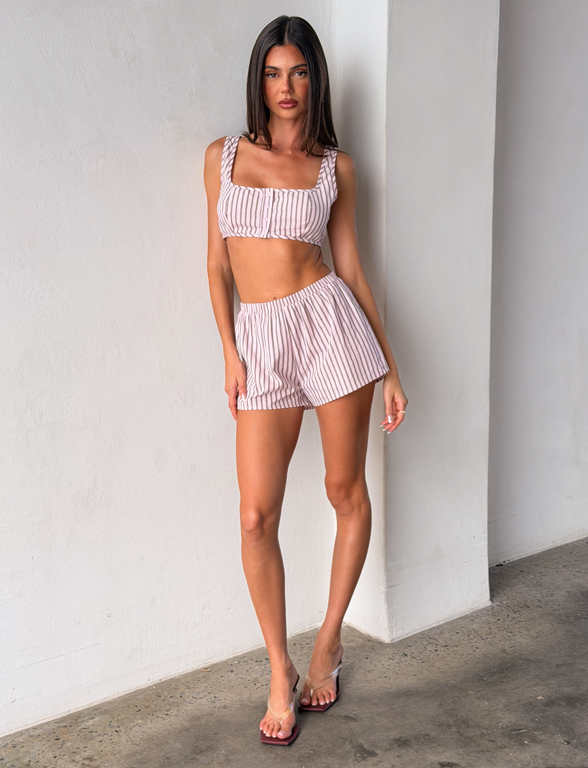 A woman with straight brown hair stands against a white wall, wearing TIGERMIST’s EZRA SHORT - PINK STRIPE set with a matching crop top and clear strap sandals, creating an easygoing indoor look.