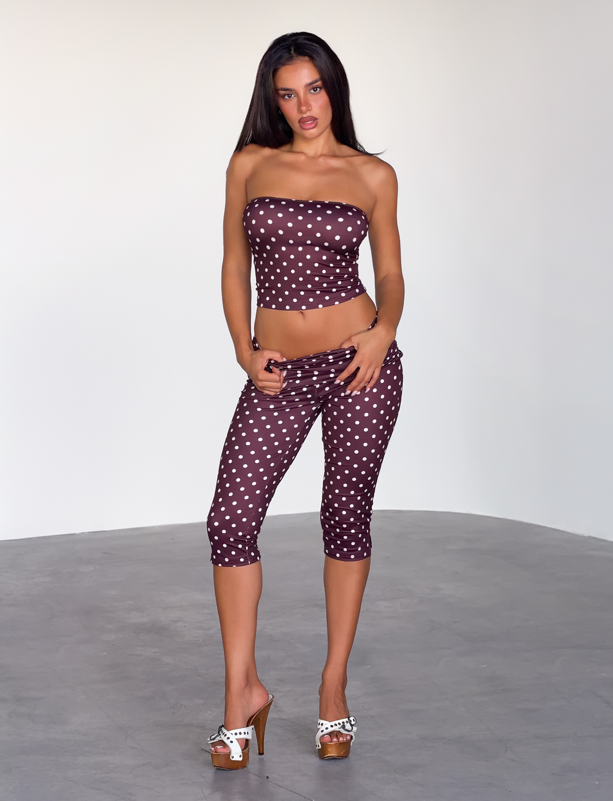 A woman stands indoors by a plain light wall, wearing the TIGERMIST VALI POLKA DOT TOP in chocolate pink dot and matching capri pants, paired with high-heeled sandals with bows.