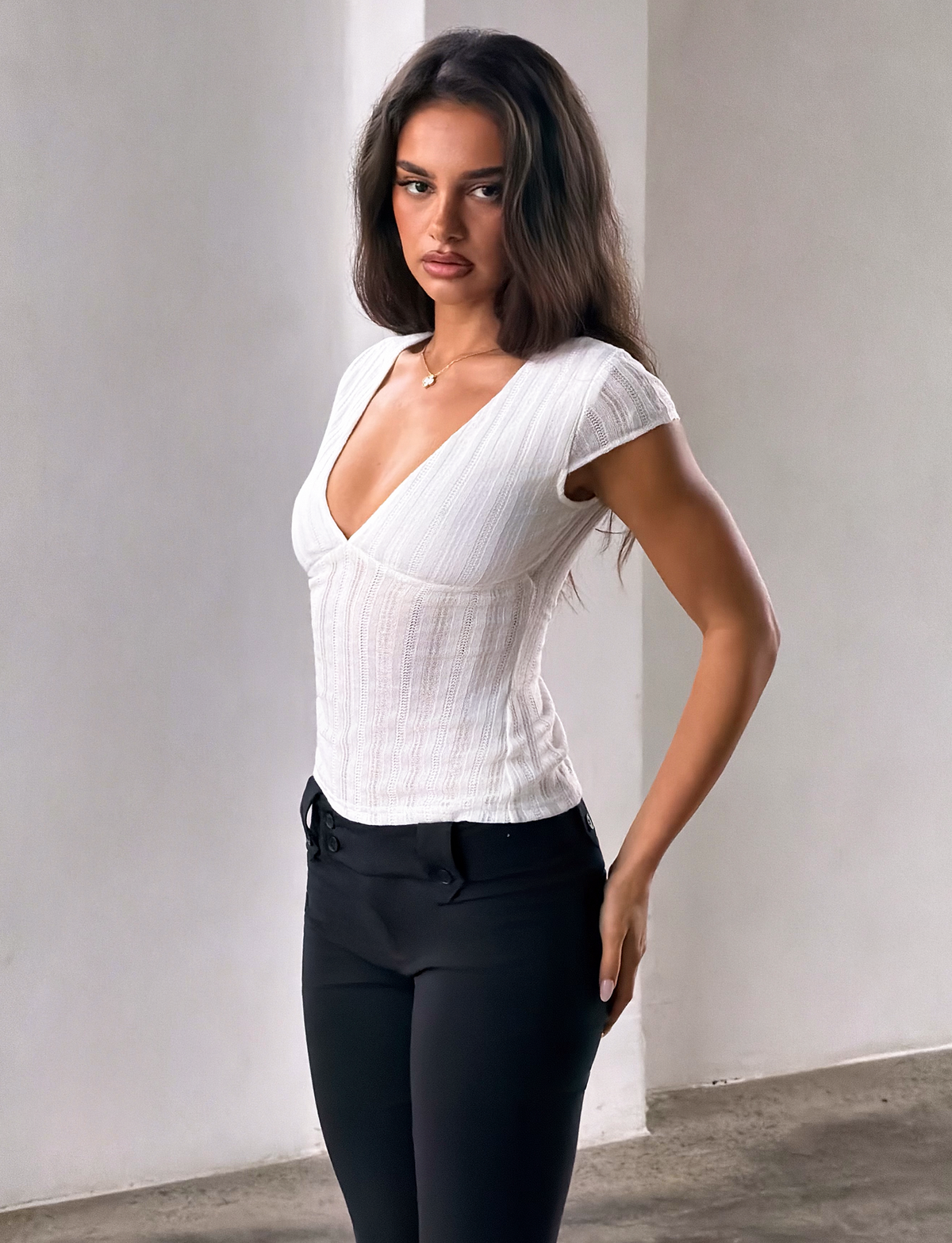 A woman with long brown hair is standing indoors near a white wall, looking at the camera with a neutral expression. She is wearing the TIGERMIST LONA TOP in white, styled with black pants.