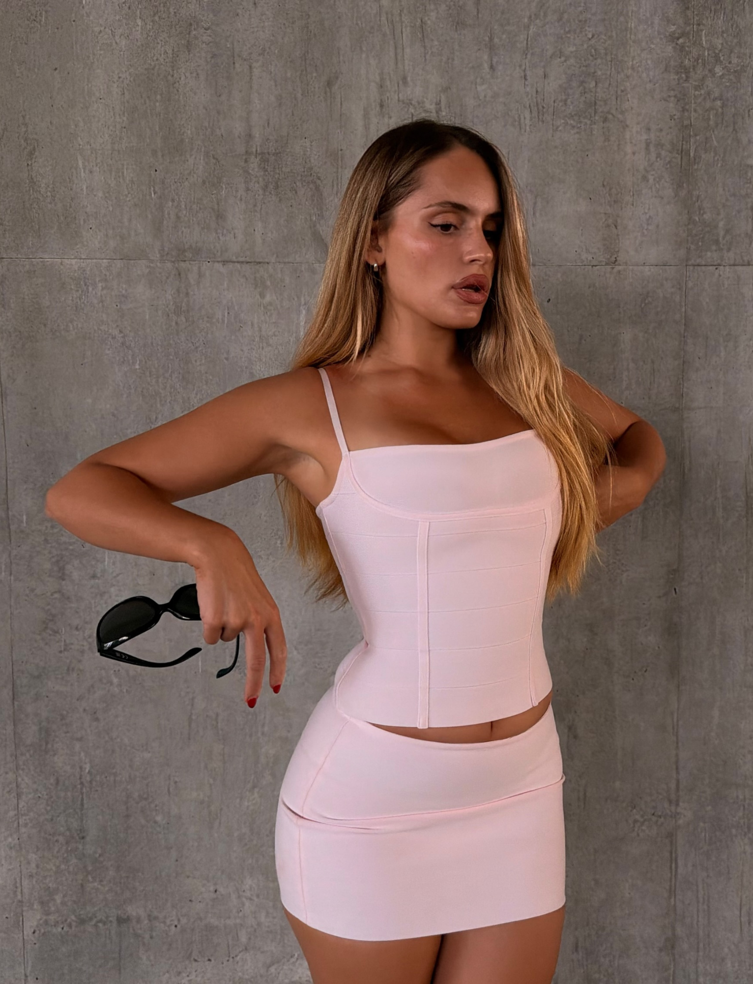 A woman with long blonde hair poses in a fitted light pink top and the TIGERMIST TAHNIA BANDAGE MINI SKIRT in Ballet Pink, crafted from sculpting bandage material. She holds black sunglasses in one hand while standing against a textured gray concrete wall.