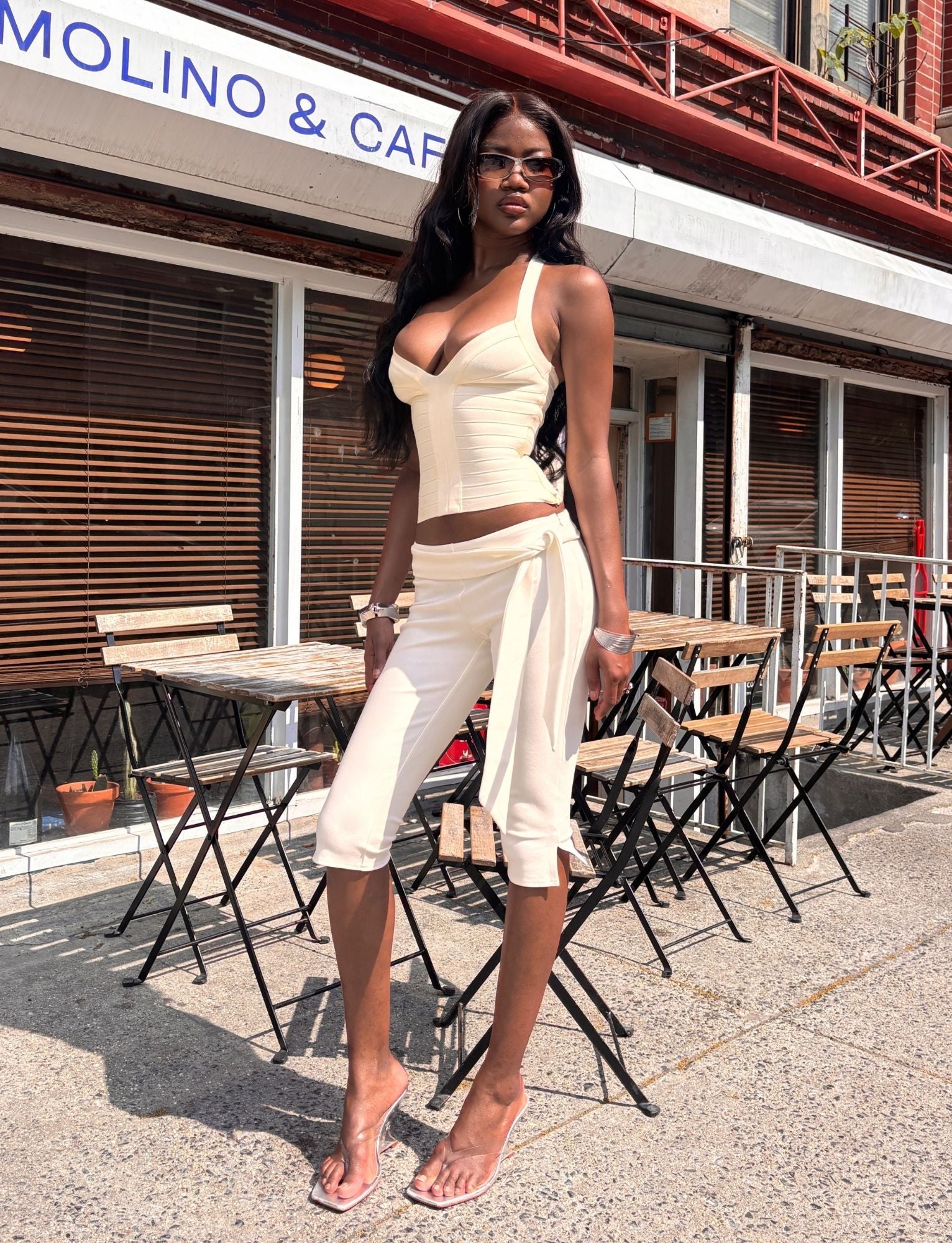 Wearing the TIGERMIST PILAR CAPRI - WHITE, a woman in a chic cream ensemble with slim leg white ponte capri pants featuring a removable waist tie and clear heels stands confidently on a sunny sidewalk outside a café with wooden tables, chairs, and large windows adorned with blinds.