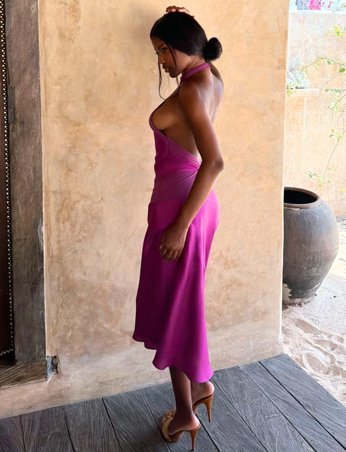 A woman in the TIGERMIST ZAYA MIDI DRESS - MULBERRY and high heels stands sideways indoors, hair in a low bun, looking down against a neutral background.