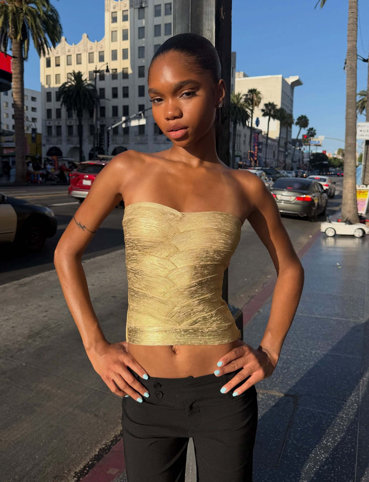 Wearing the TIGERMIST NIYA METALLIC BANDAGE TOP in gold, a person stands confidently on a city sidewalk in black pants, with palm trees, cars, and tall buildings in the background.